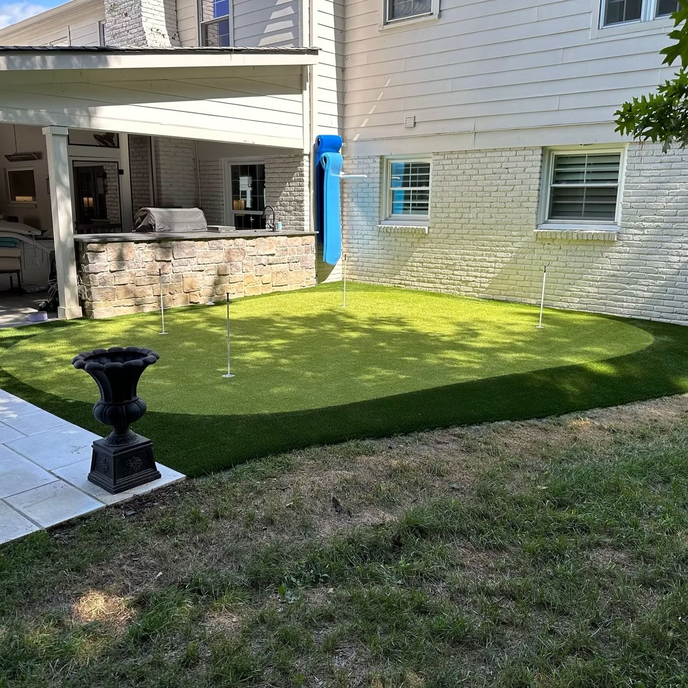 Backyard with putting green, bordered by grass, next to a white brick house with a covered patio.