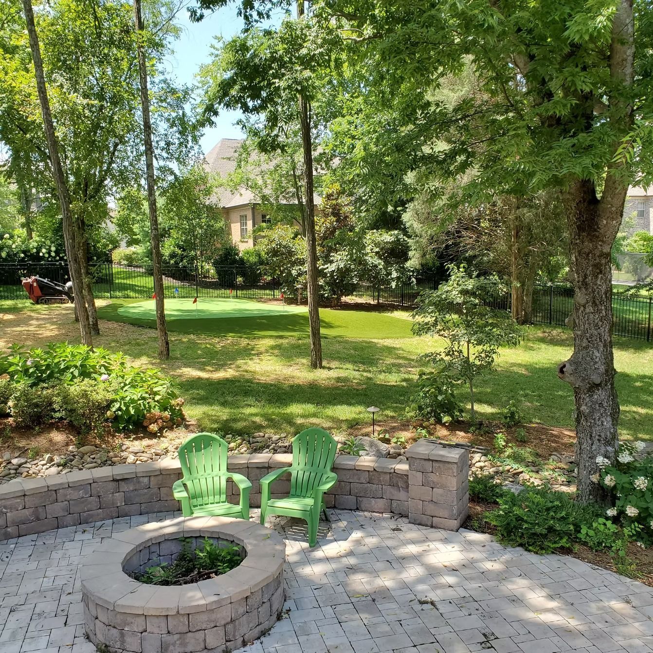 Patio with green chairs, fire pit, and brick wall. Trees and lawn in the background. Sunny day.