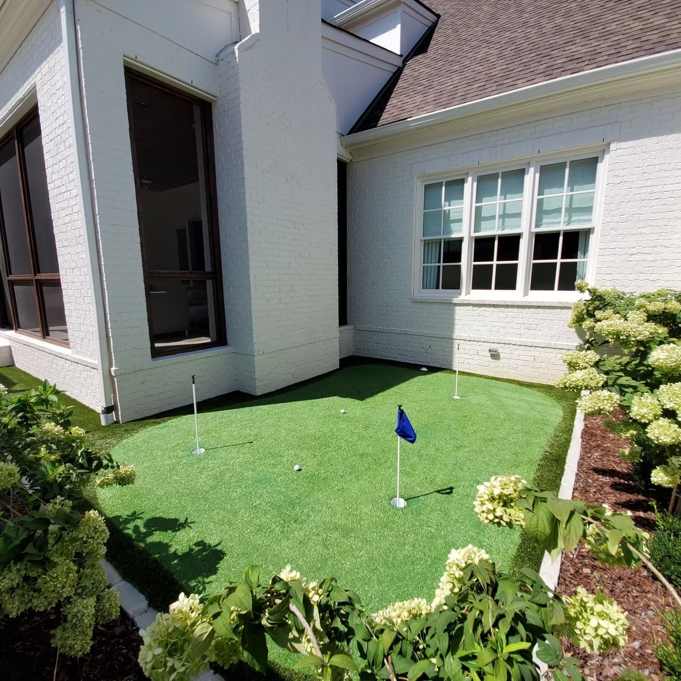 Small backyard putting green with white house and landscaping.