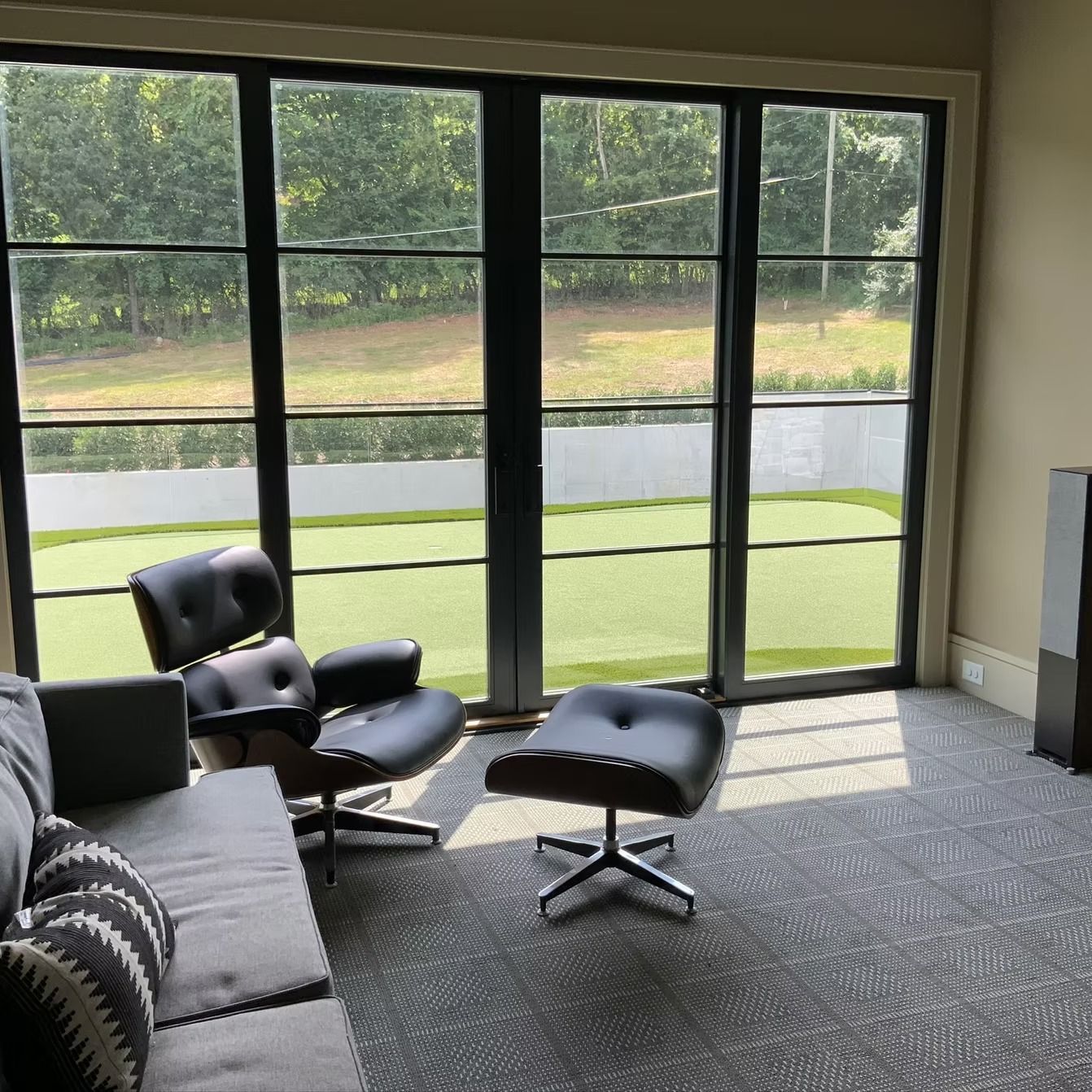 A modern living space with black-framed windows overlooking a green lawn. Includes a leather chair, ottoman, and gray sofa.