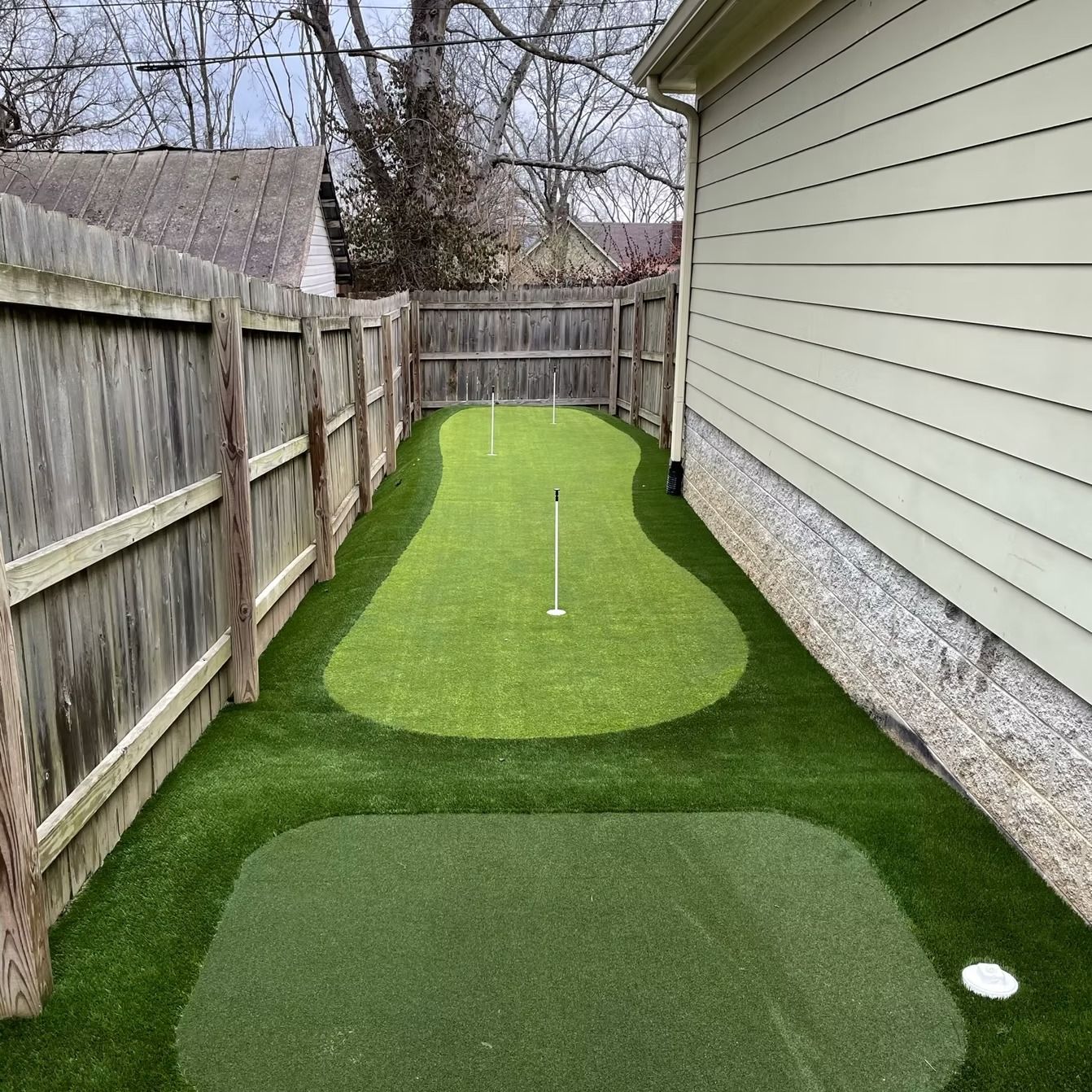 Backyard putting green, enclosed by fences and a house. Green artificial turf with golf flagpoles.