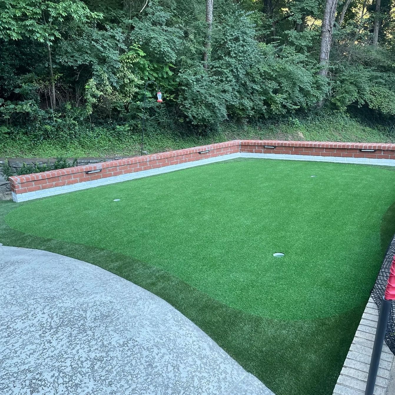 Green artificial turf putting green with brick border, next to patio, surrounded by trees.