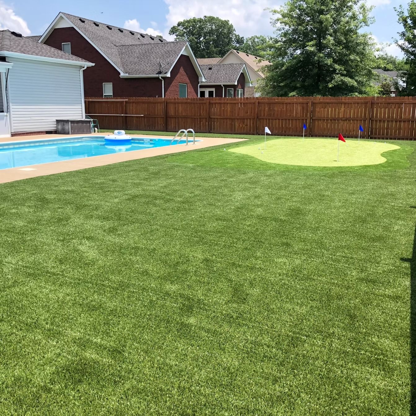 Backyard with pool, putting green, and wooden fence. Lush green lawn, blue sky, and residential houses.
