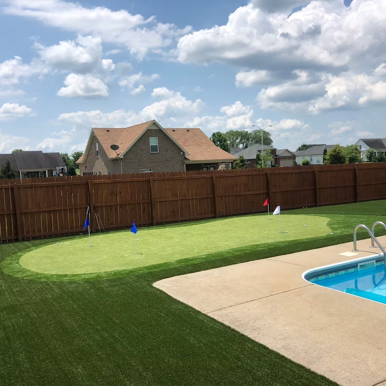 Backyard with a putting green, swimming pool, and a brown fence. Blue and white flags are visible.