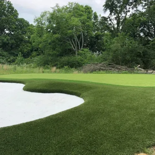 A golf green with artificial turf surrounds a white sand bunker, backed by trees.