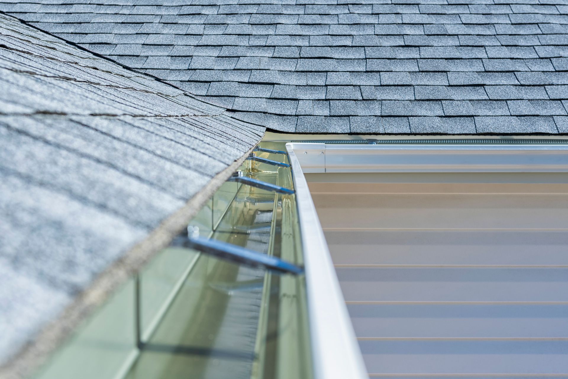 Close-up of a roof with gray shingles, gutters, and white siding.