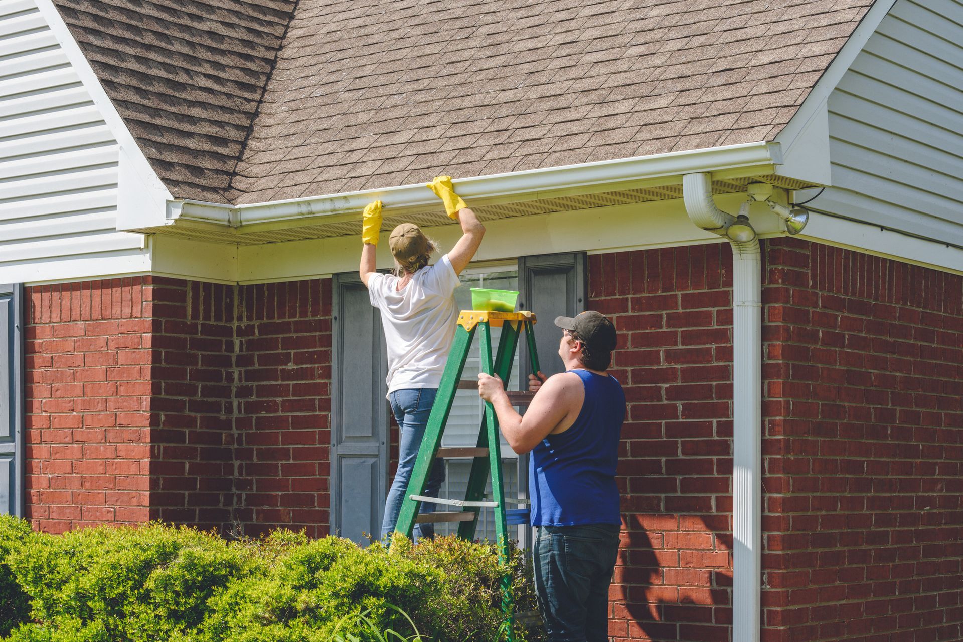 Two people cleaning gutters of a brick house using a ladder and sponges.