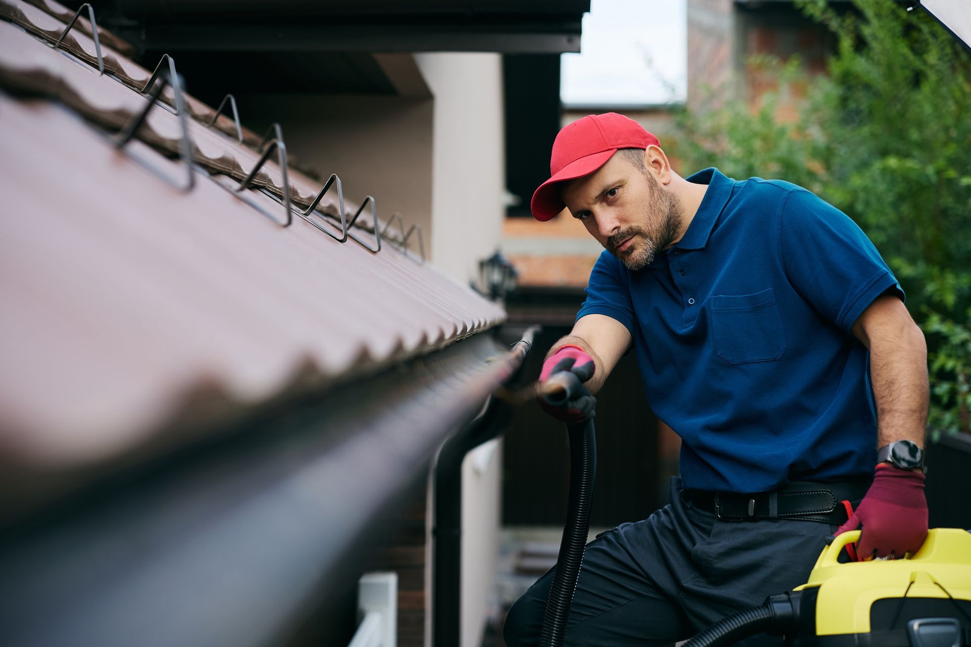Man cleaning gutters with a vacuum, wearing a red hat, blue shirt, and red gloves.