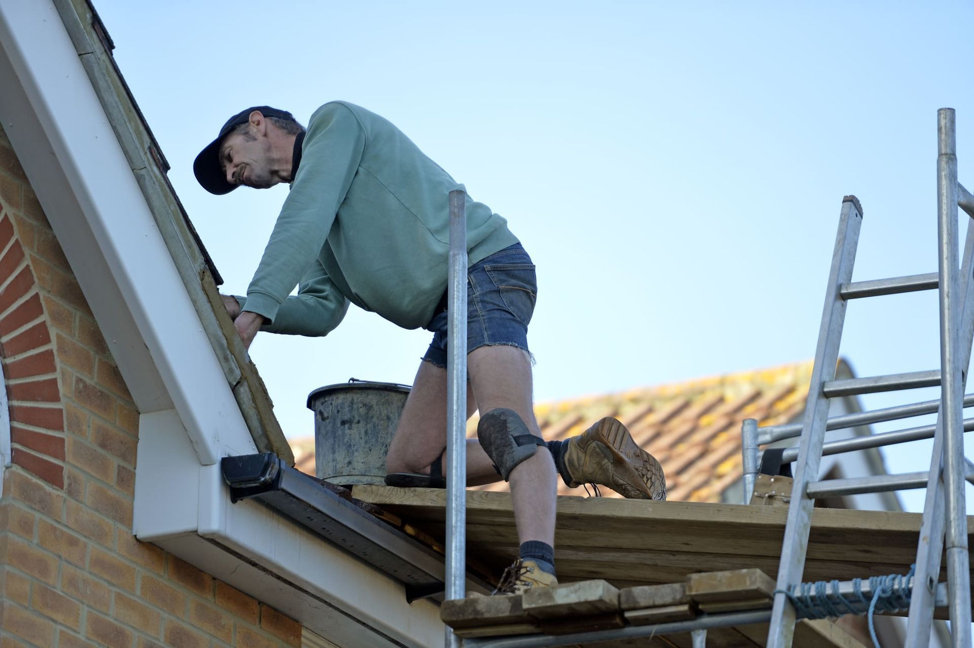 Man on scaffold repairing roof, wearing cap, light green shirt, denim shorts, and work boots.