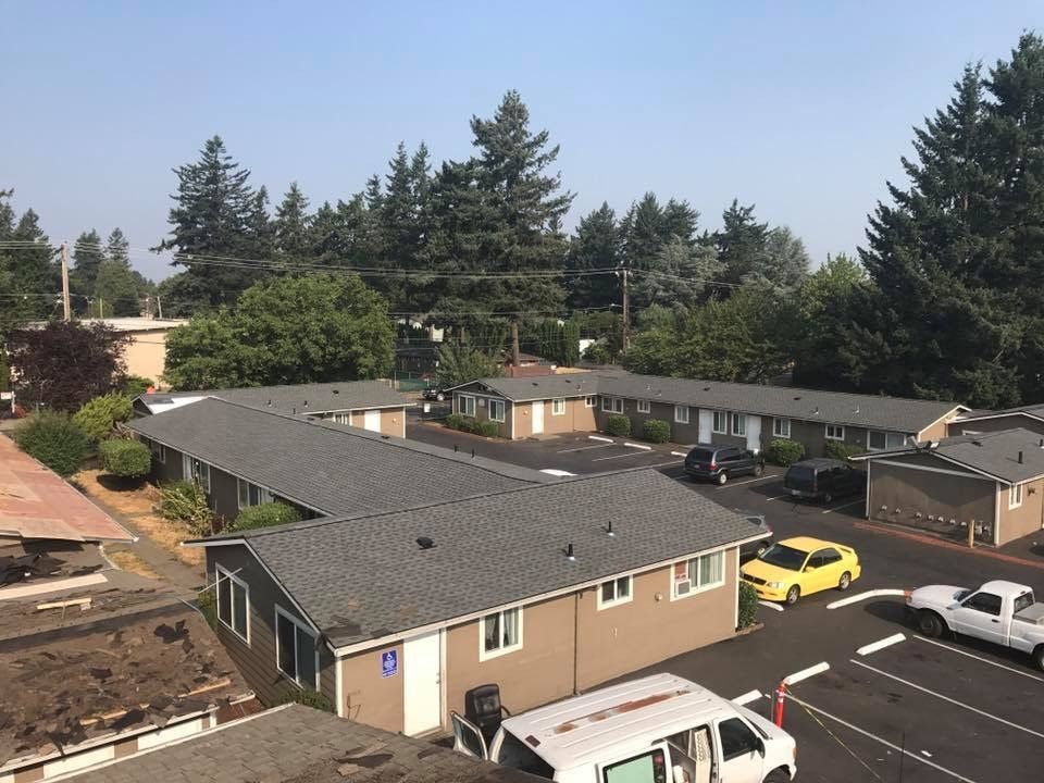 Apartment complex with gray roofs, tan walls, and parked cars, surrounded by trees on a sunny day.
