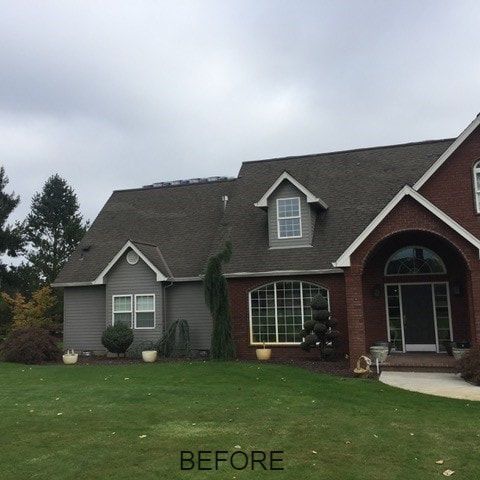 Gray and brick home with gray siding, dormers, and a large front lawn. Overcast sky.