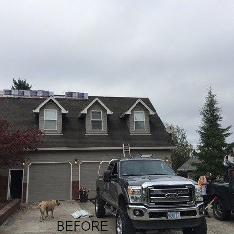 House with gray siding, three dormers, and a truck in the driveway. Roofing materials on the roof. Cloudy sky.
