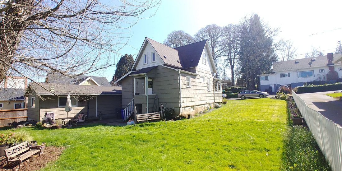 House with green lawn, white fence, and surrounding trees on a sunny day.