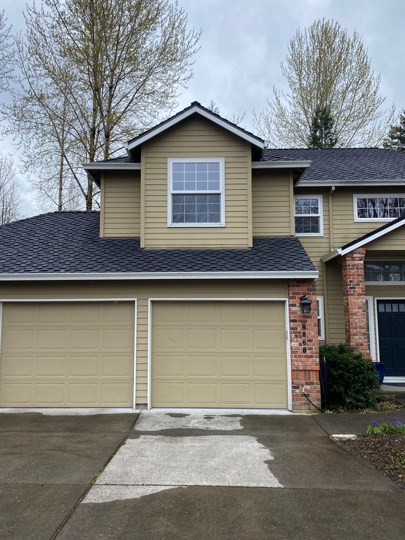 Two-story beige house with two garage doors, black roof, and brick accents. Paved driveway, trees in the background.