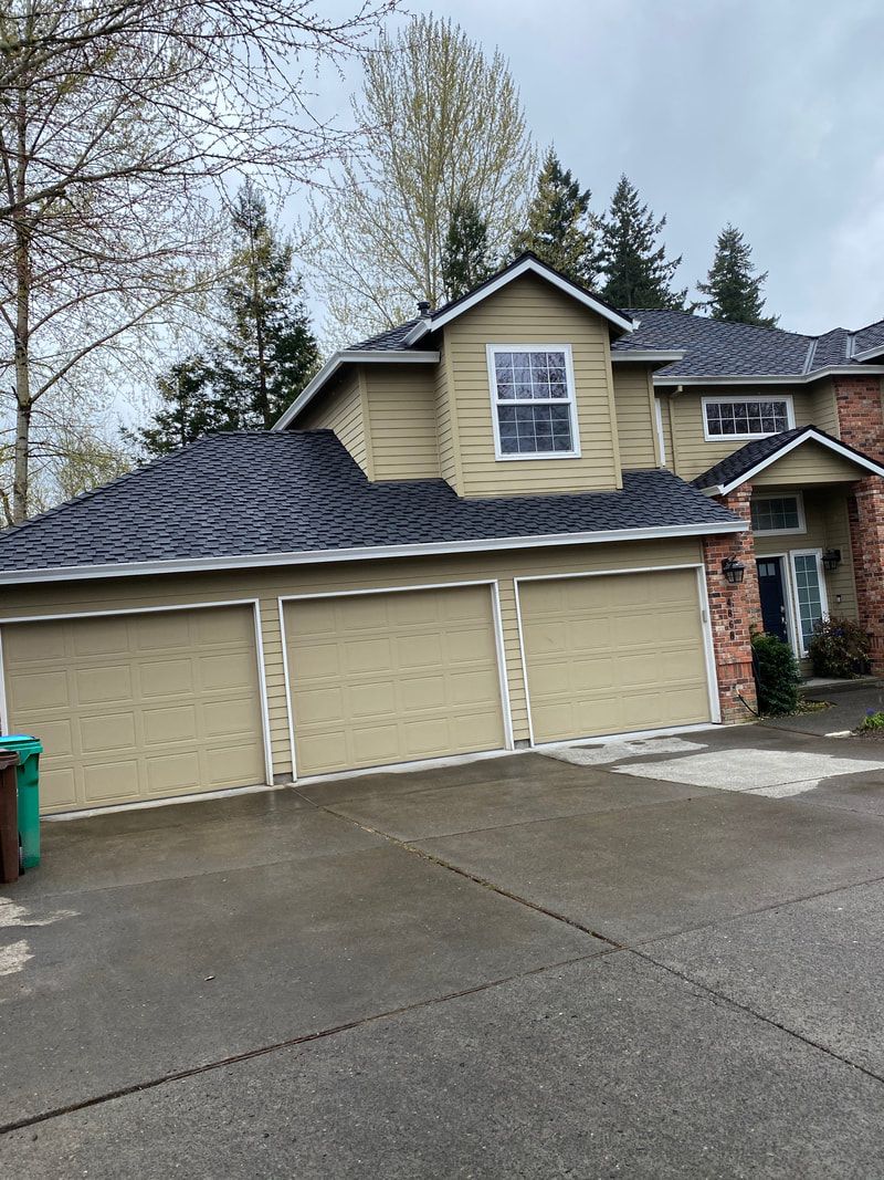 Two-story beige house with three garage doors, dark roof, brick accents, and a wet driveway.