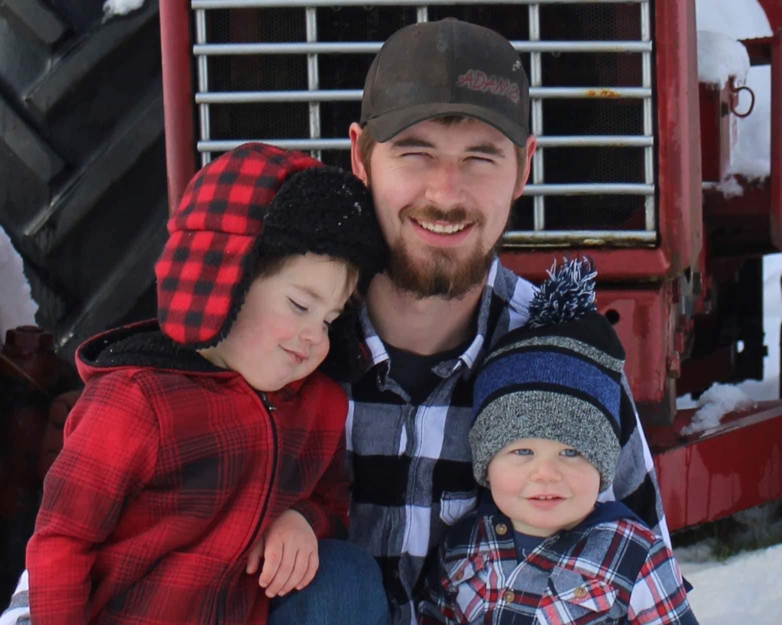 Man with two young children in winter attire smiles in front of a red tractor.