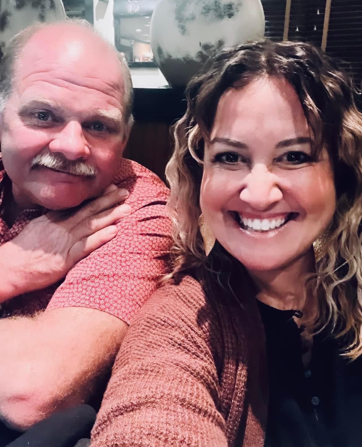 Man with mustache and woman smiling for a selfie, indoors. Man in red shirt, woman in brown sweater.