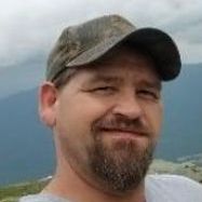 Man wearing camouflage cap smiles; outdoor setting with mountains.