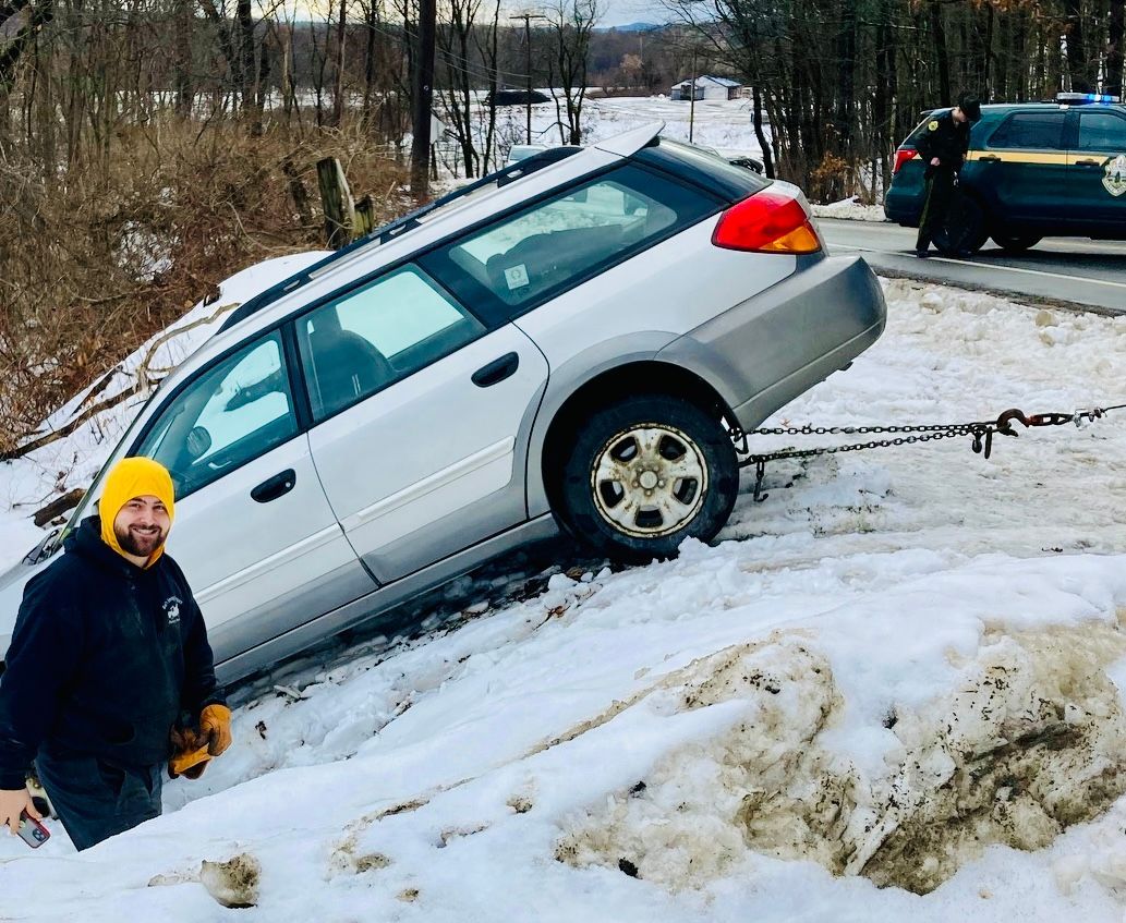 Man by a silver Subaru wagon stuck in snow bank, being pulled out by a tow truck on roadside.