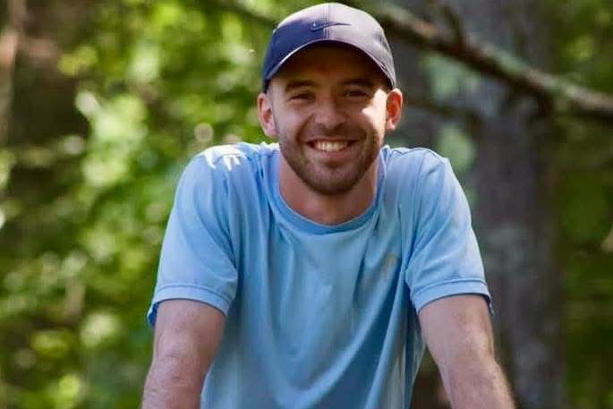 Man with a light blue shirt and a dark blue cap smiles outdoors in front of blurred greenery.