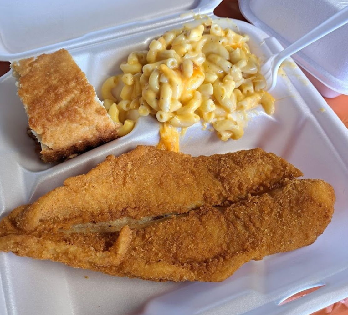 A styrofoam container filled with fried fish macaroni and cheese and cornbread