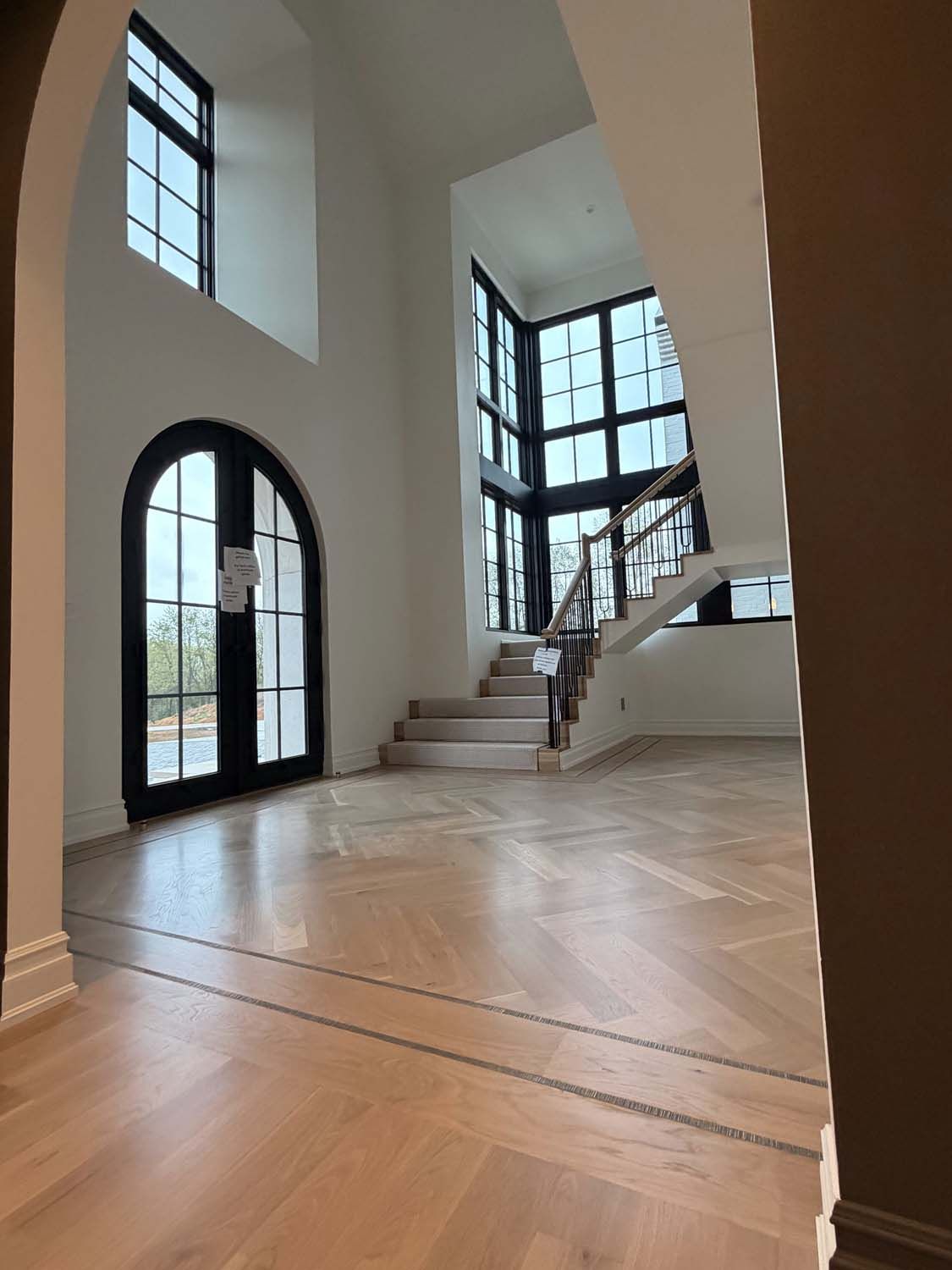 A spacious, bright foyer with white walls, herringbone wood floors, a grand arched entrance, and an open staircase.