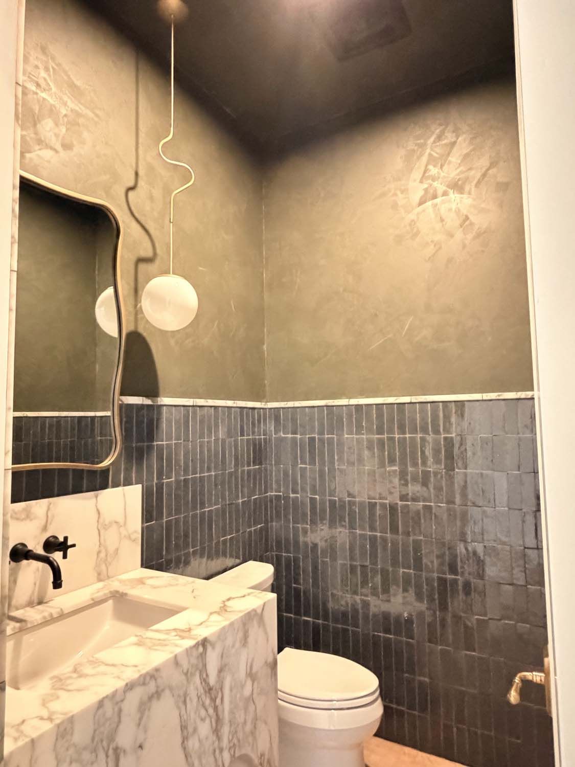 A modern powder room with a white marble vanity, dark blue vertical wall tiles, grey textured walls, and pendant lighting.