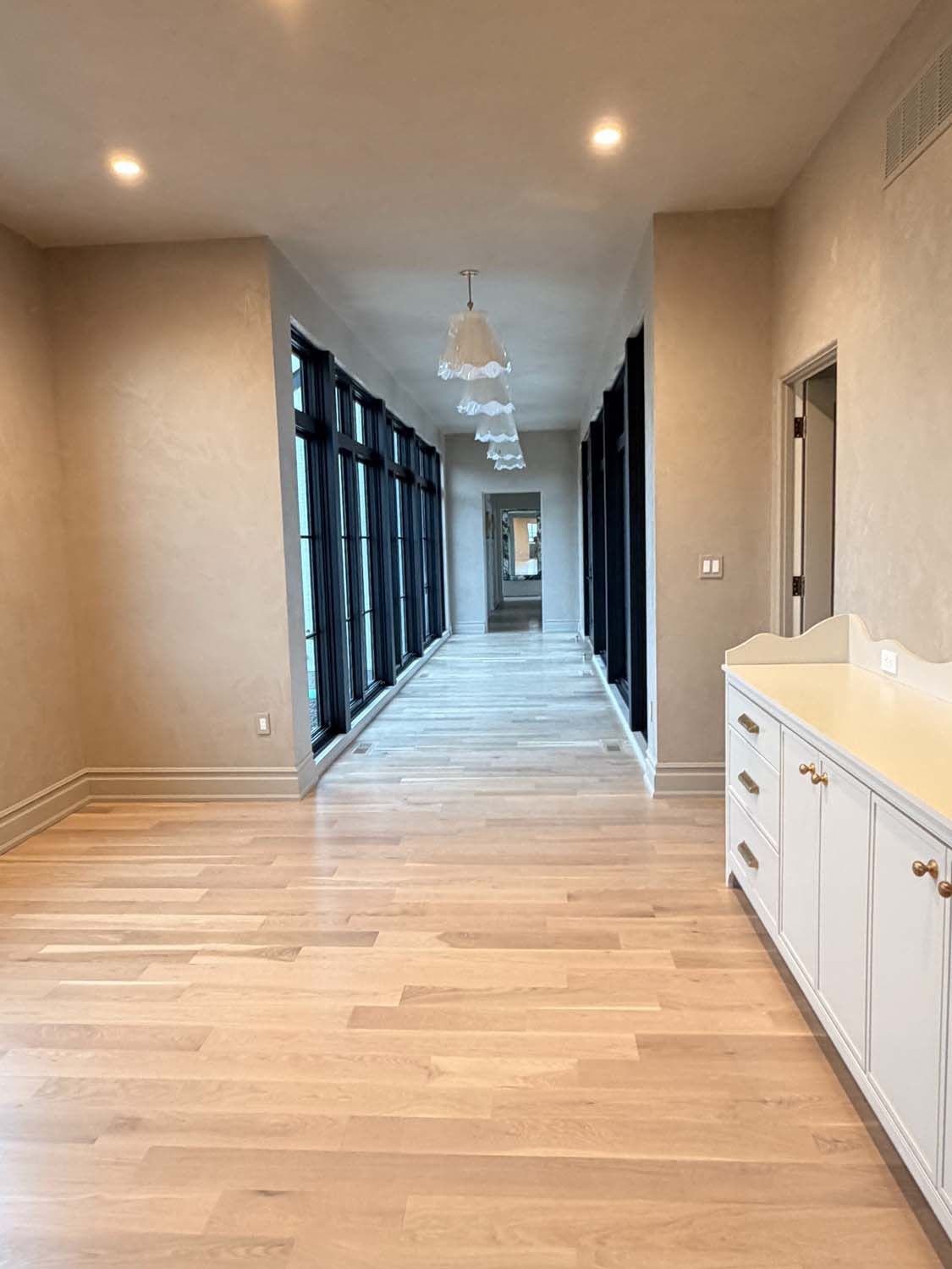 A hallway with light wood floors and beige walls, featuring floor-to-ceiling windows with dark curtains and white cabinets.