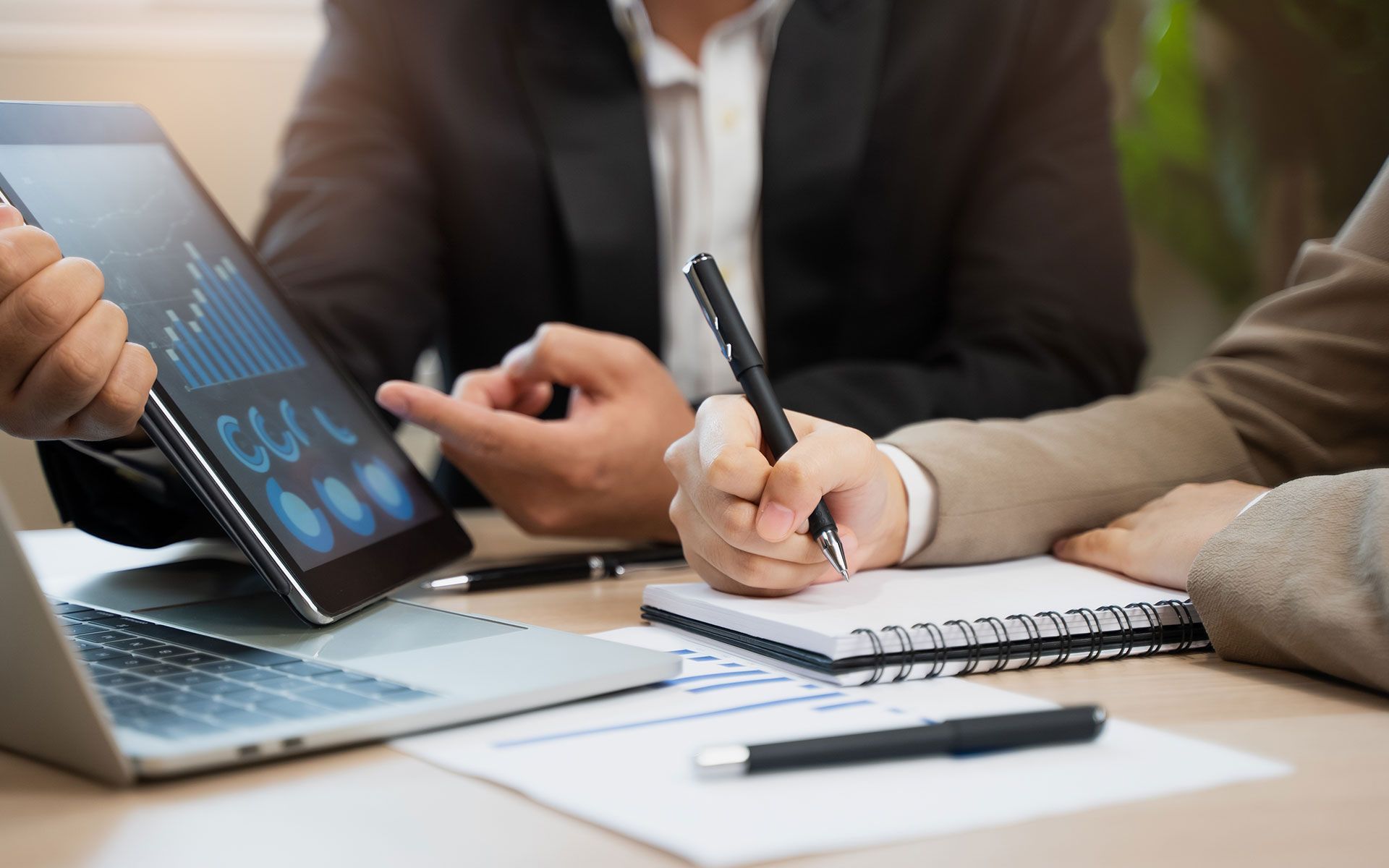 Businesspeople at a table, reviewing data on a tablet and taking notes in a meeting.