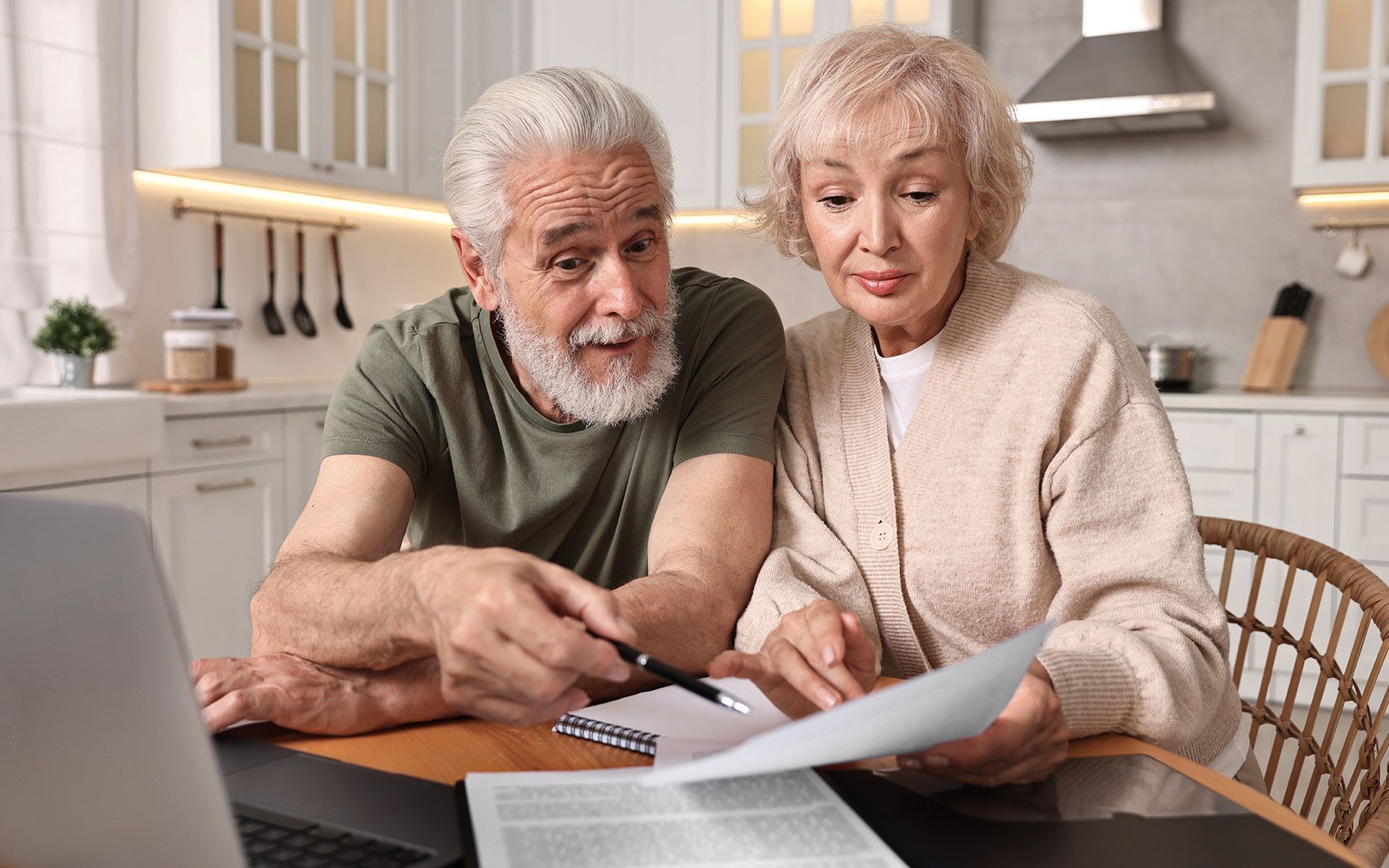Senior couple reviewing paperwork at a table, looking at documents with concerned expressions in a kitchen.