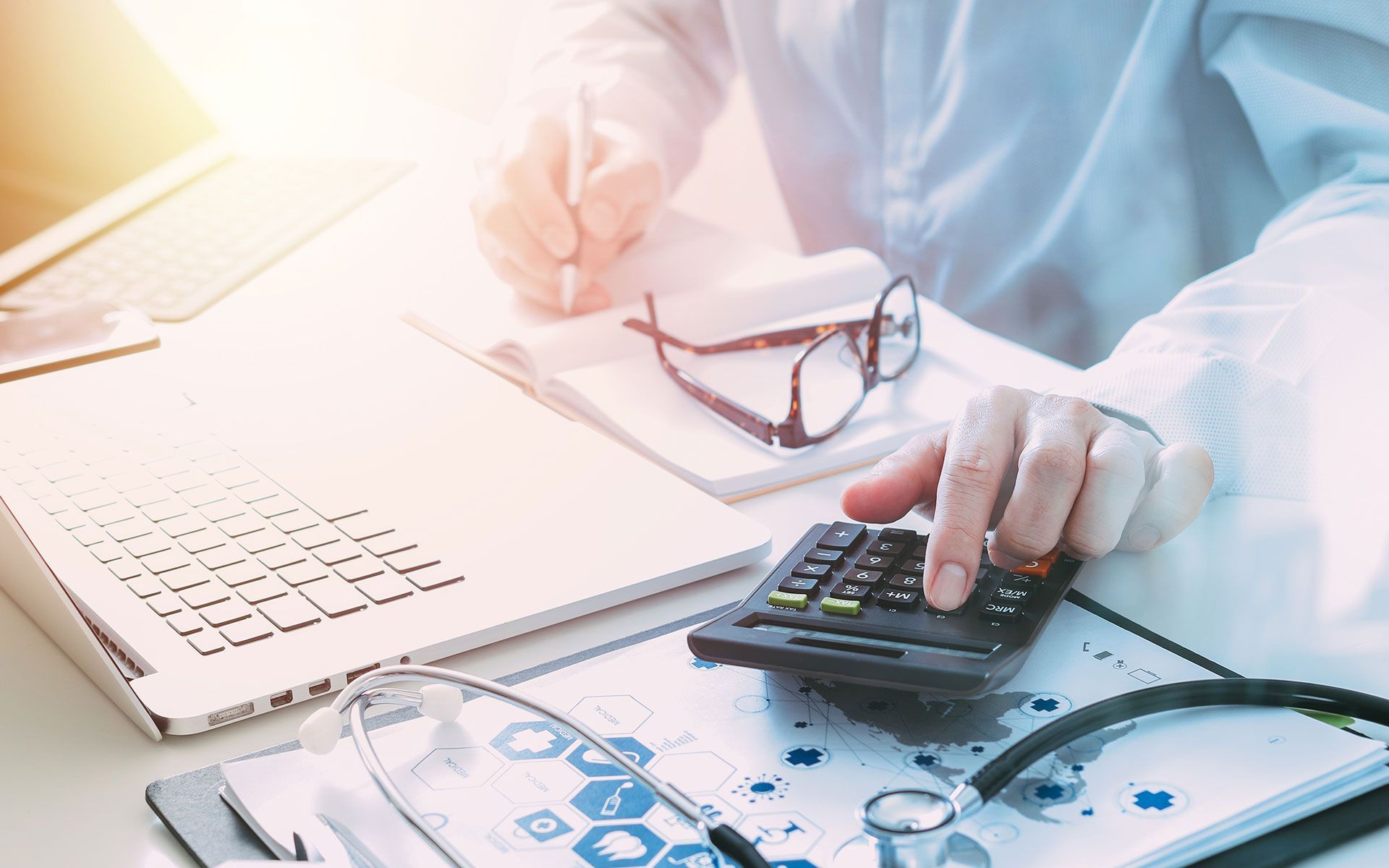 A doctor at a desk, calculating with a calculator, and taking notes, with a laptop and stethoscope.