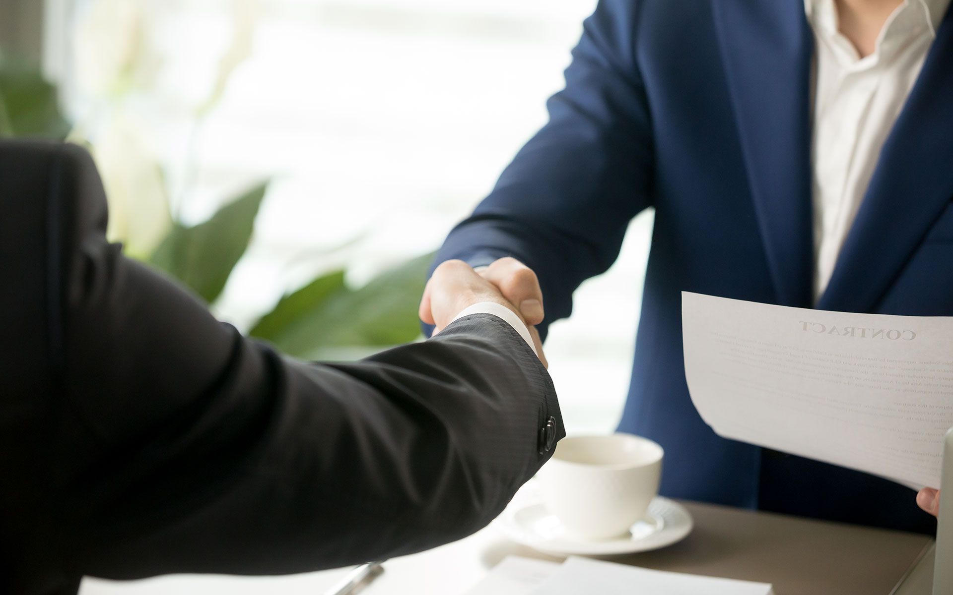Two businessmen shaking hands; blue and black suits, bright room, documents and coffee.