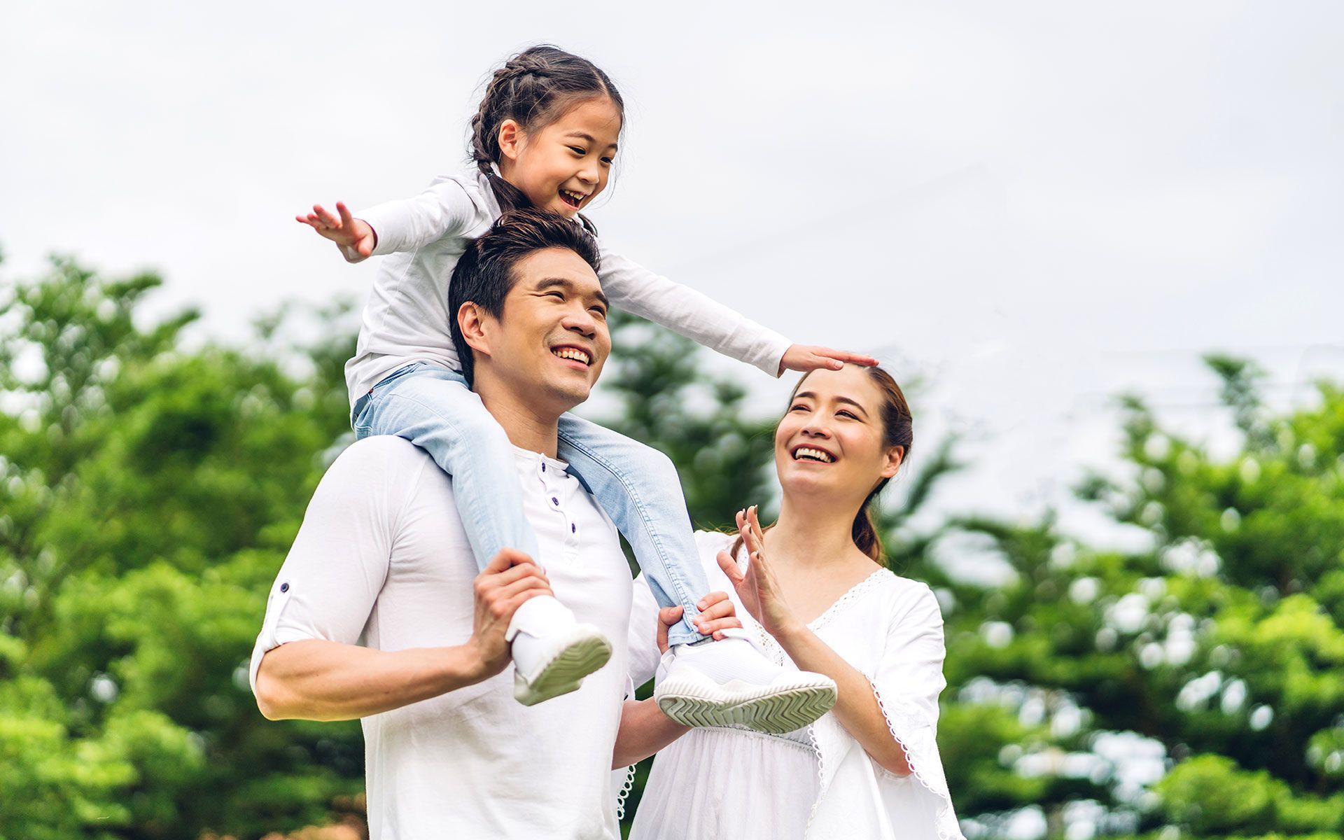 Asian family of three outdoors, smiling: father carrying daughter on shoulders, mother smiling next to them.