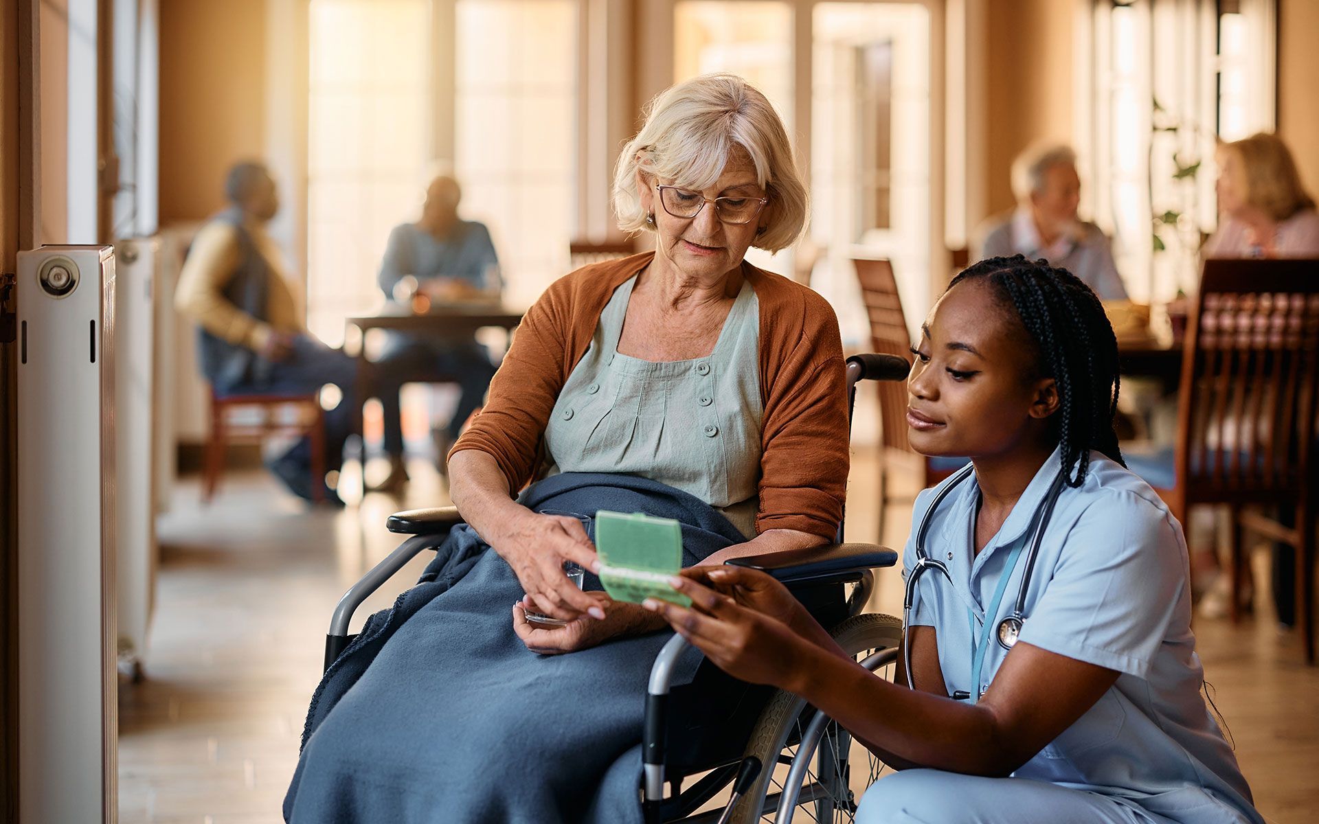 Nurse showing a medication card to an elderly woman in a wheelchair. Other residents are in the background.
