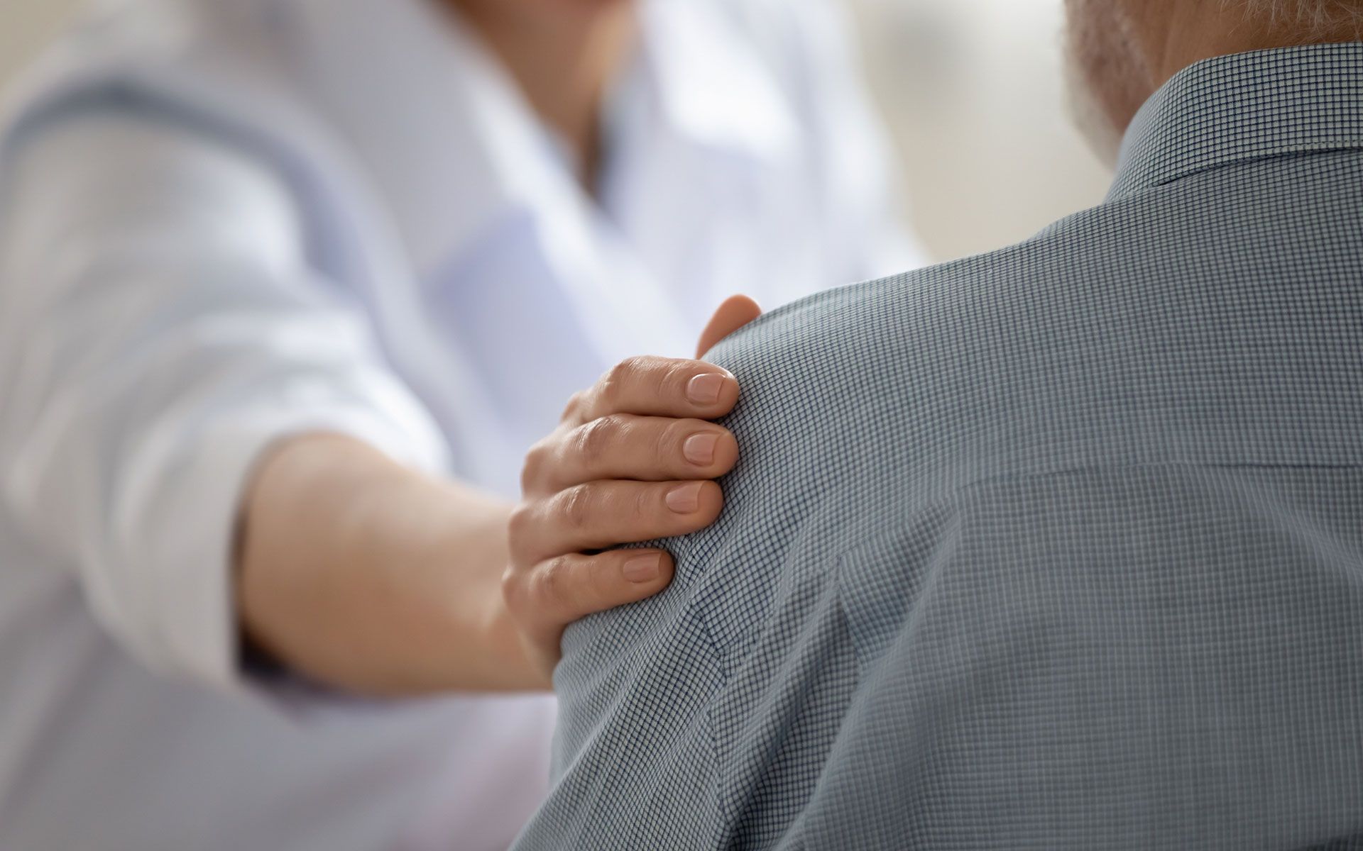 Woman's hand on man's shoulder offering support. Light clothing, indoors.