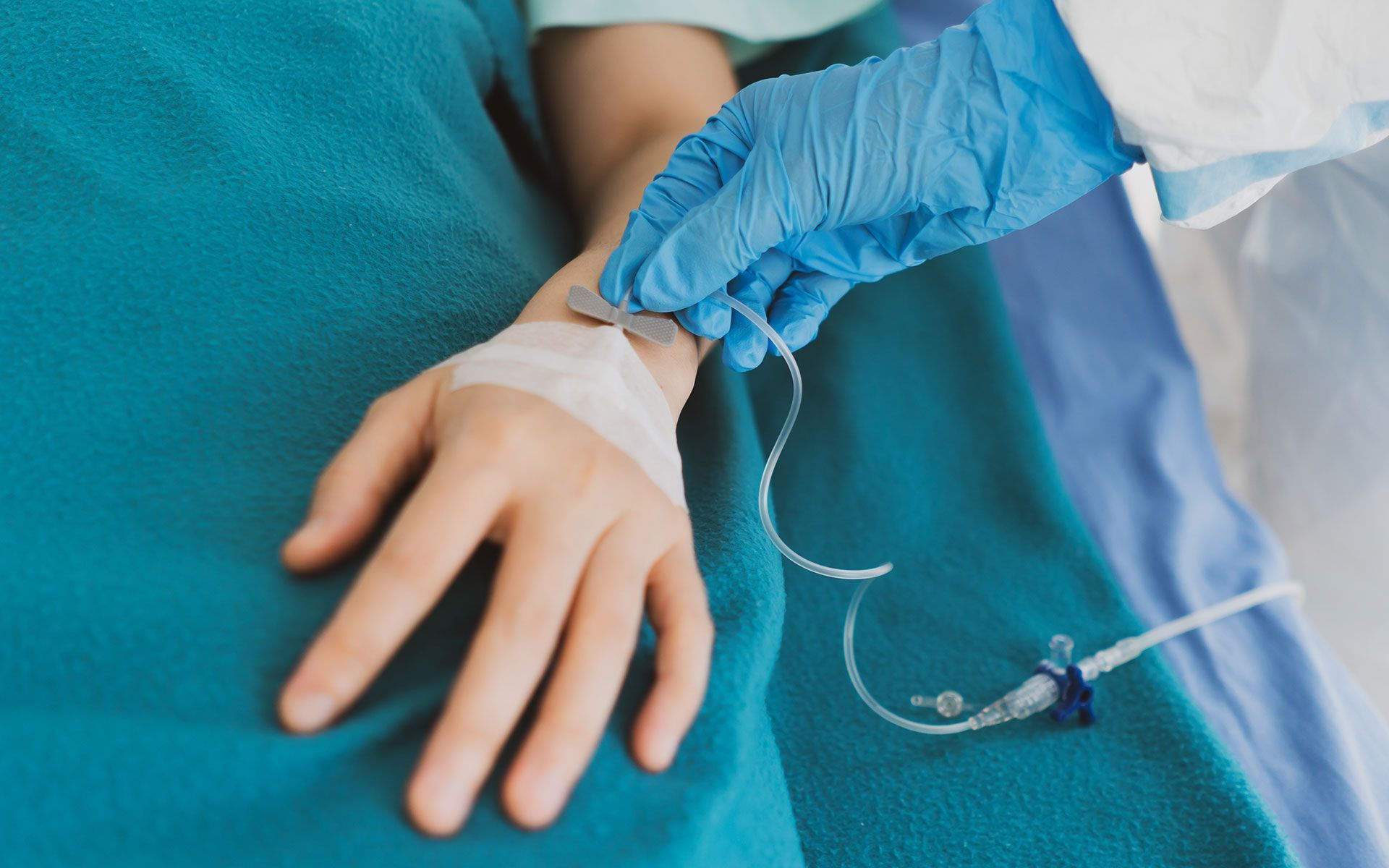 Person's hand with IV line attached, being attended to by person wearing blue gloves, in a hospital setting.
