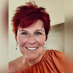 Woman with short red hair, smiling. Wearing orange top, blue earrings.