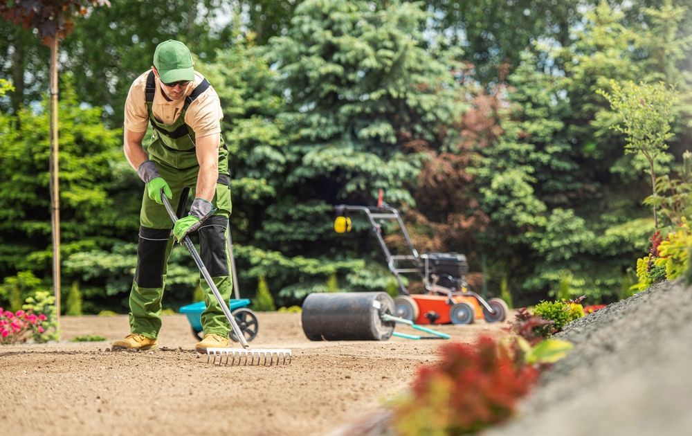 Un uomo sta rastrellando la terra in un giardino con un tosaerba sullo sfondo.