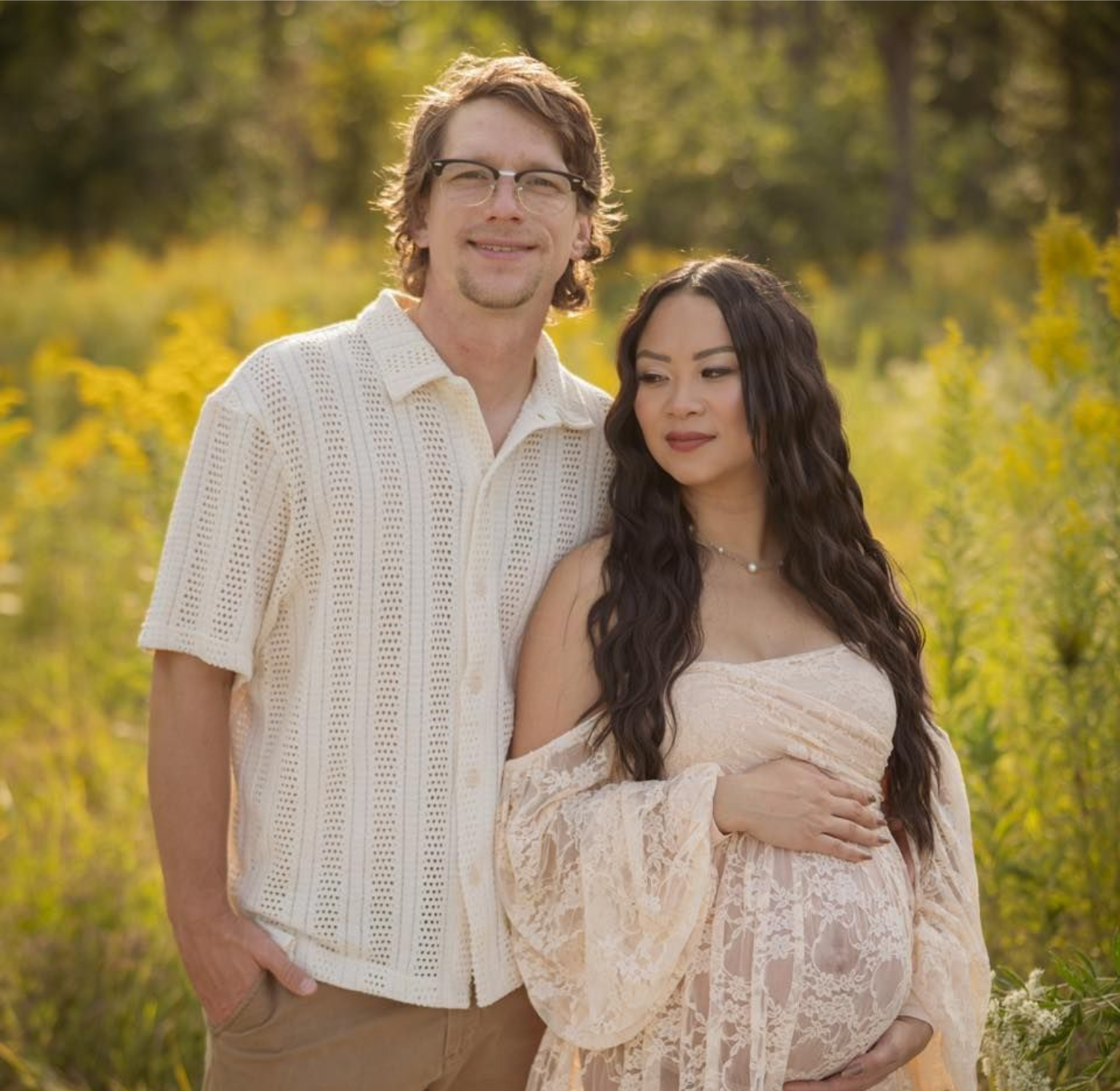Pregnant person and partner pose outdoors in field of yellow flowers.