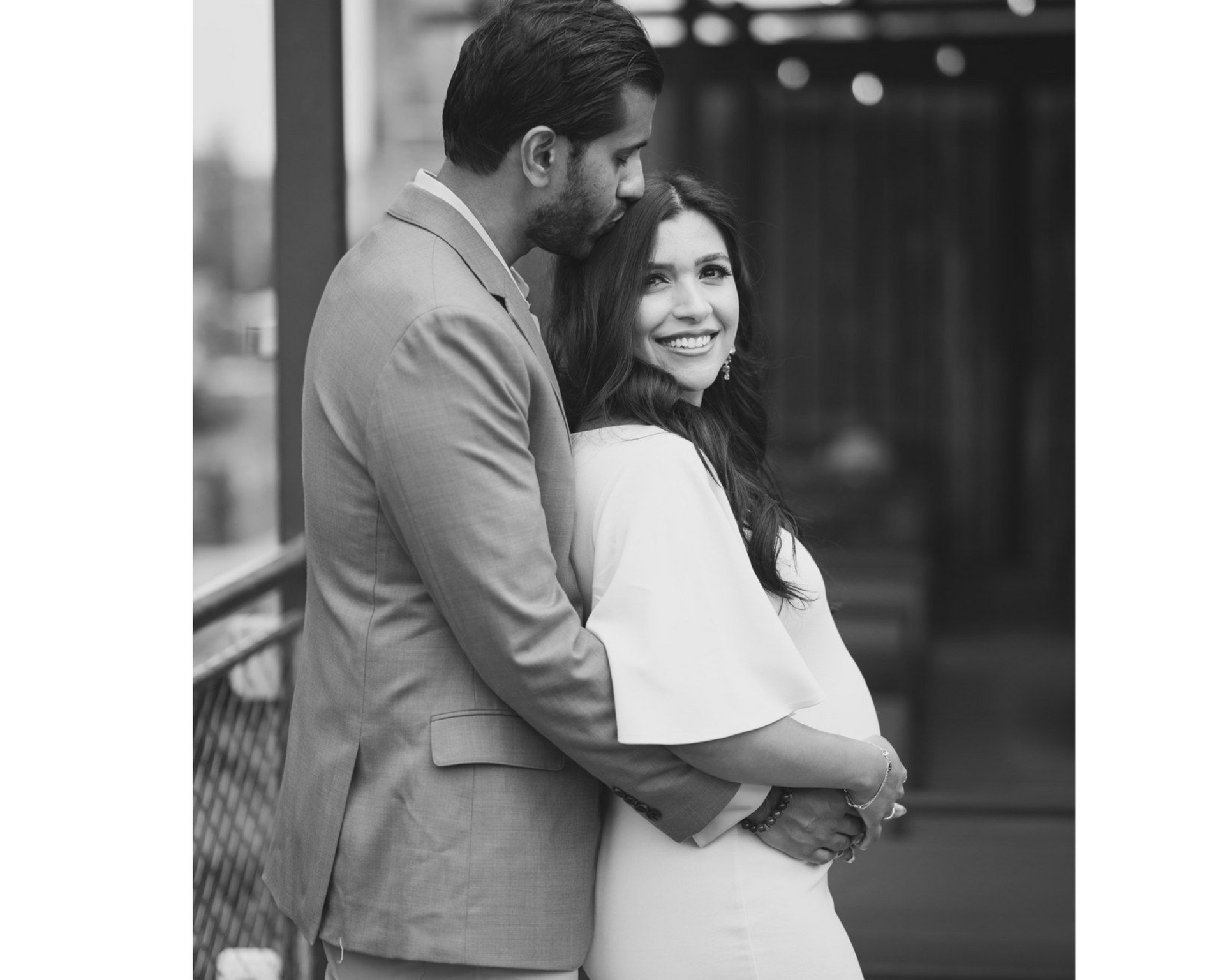 Man in a suit embraces a woman in a white dress, both smiling; outdoor setting.