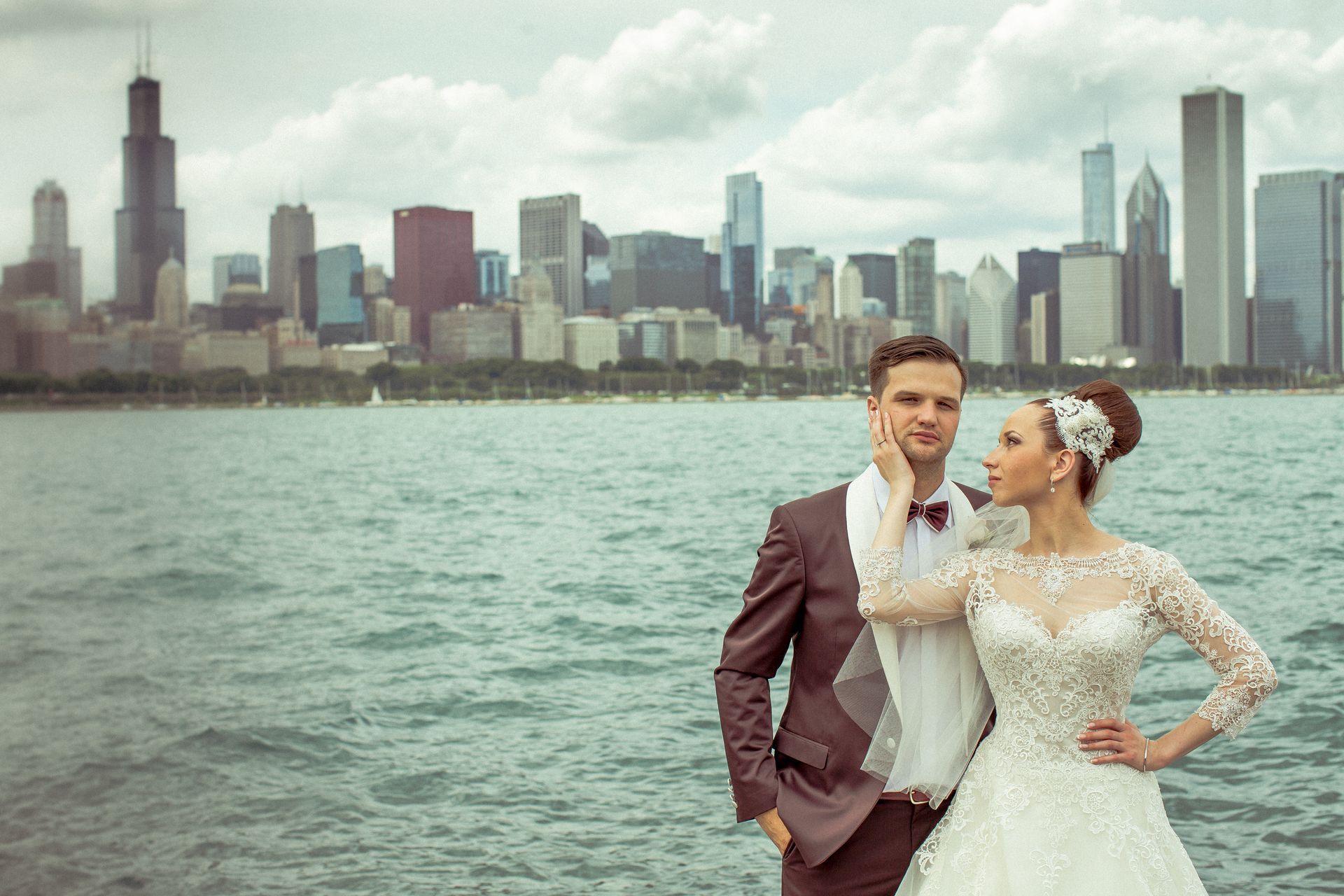 Bride and groom pose by lake with Chicago skyline in background. Woman in white gown touches man's face.