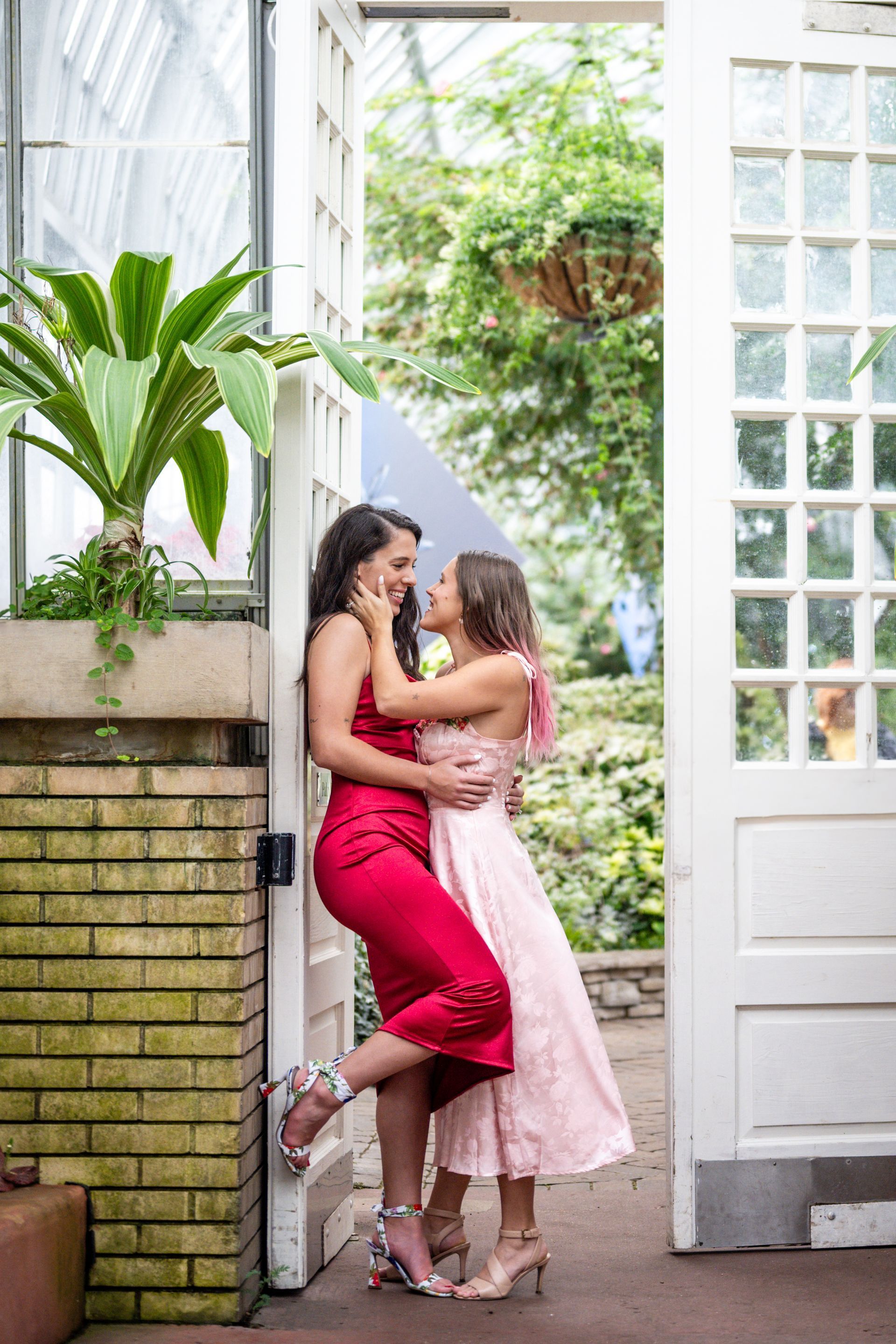Two women embrace in a greenhouse doorway. One wears a red dress, the other a pink dress.