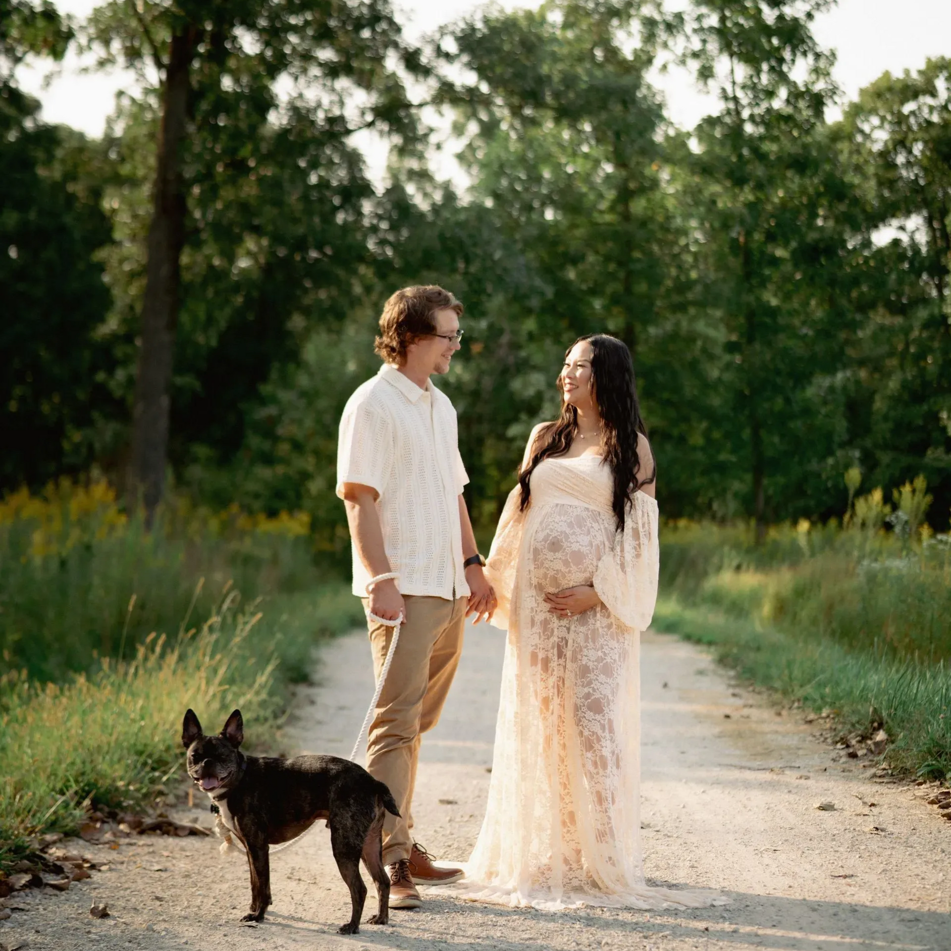 Couple on path, expecting, holding hands, dog at their feet, nature setting.