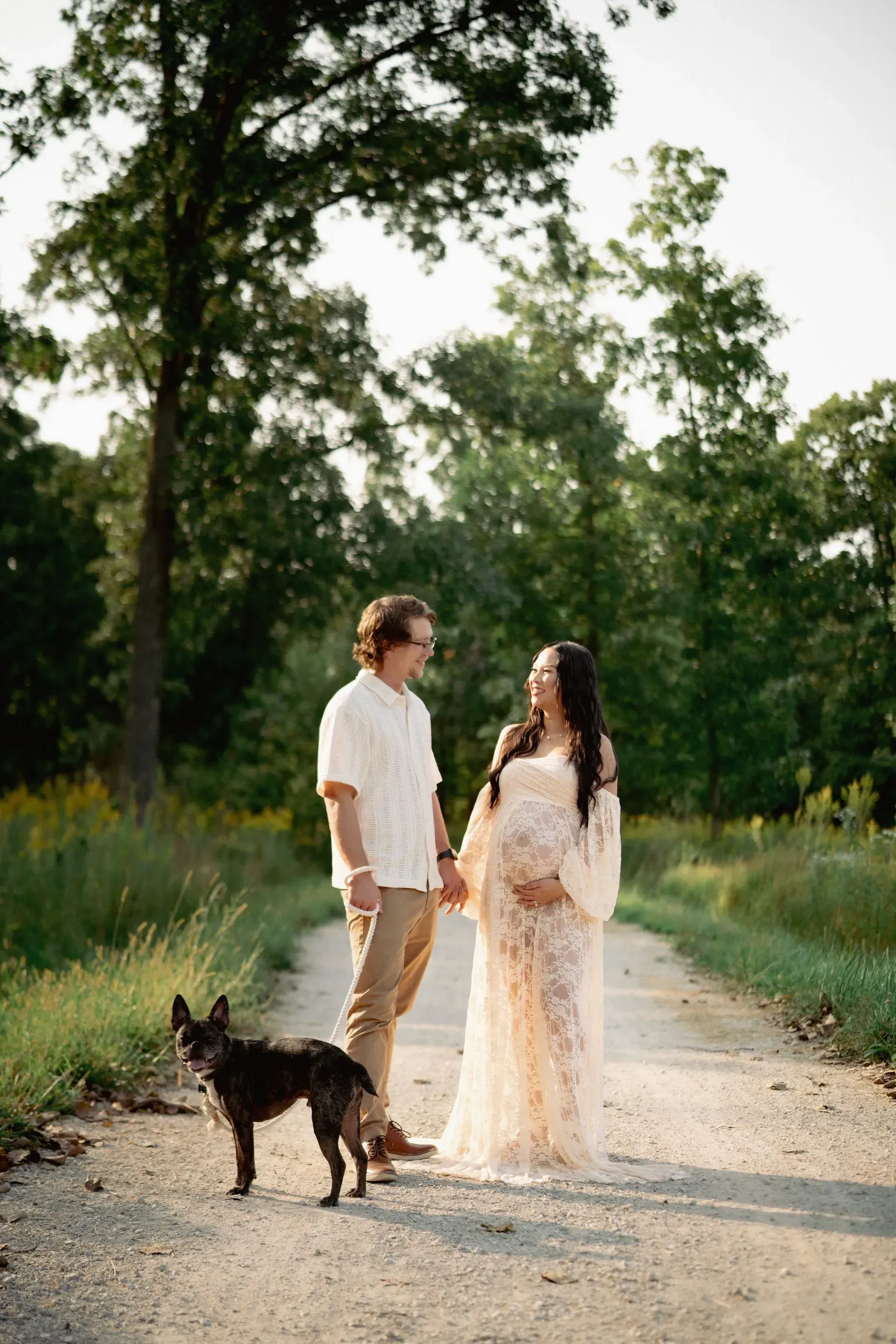 Couple holding hands on a dirt path with their dog; woman in maternity dress. Trees in background.