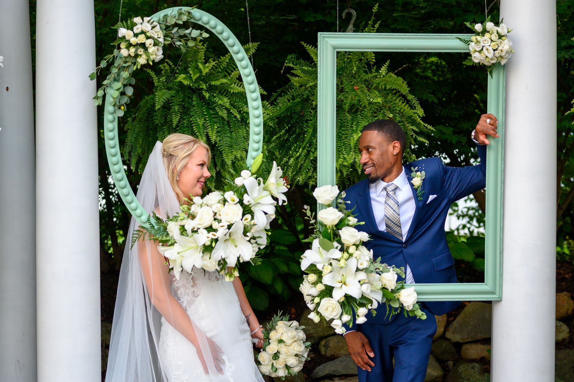 Bride and groom look at each other, holding hands, in front of a wall of framed art. The woman is wearing a white dress.
