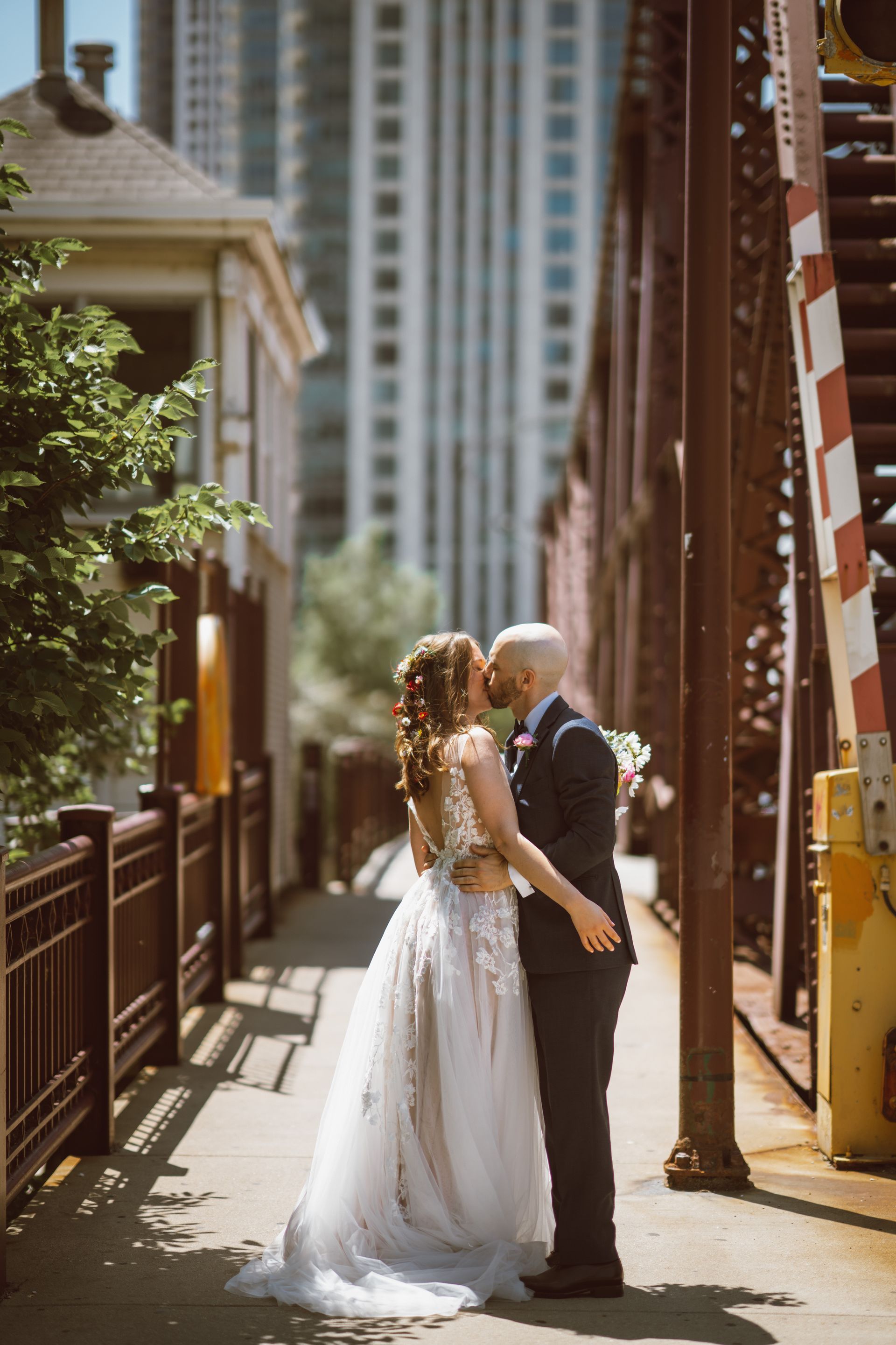 Newlyweds kiss on a bridge, groom in a suit, bride in a wedding dress. City buildings in the background.