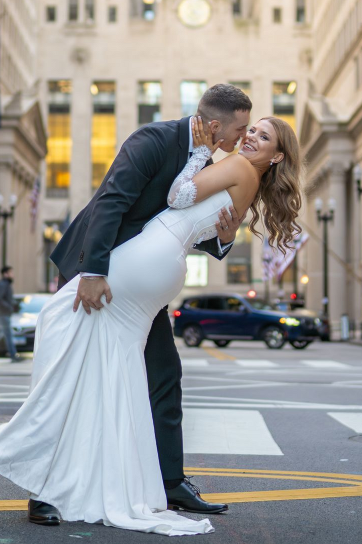 Couple in wedding attire embrace on a city street; groom leans bride back, laughing.