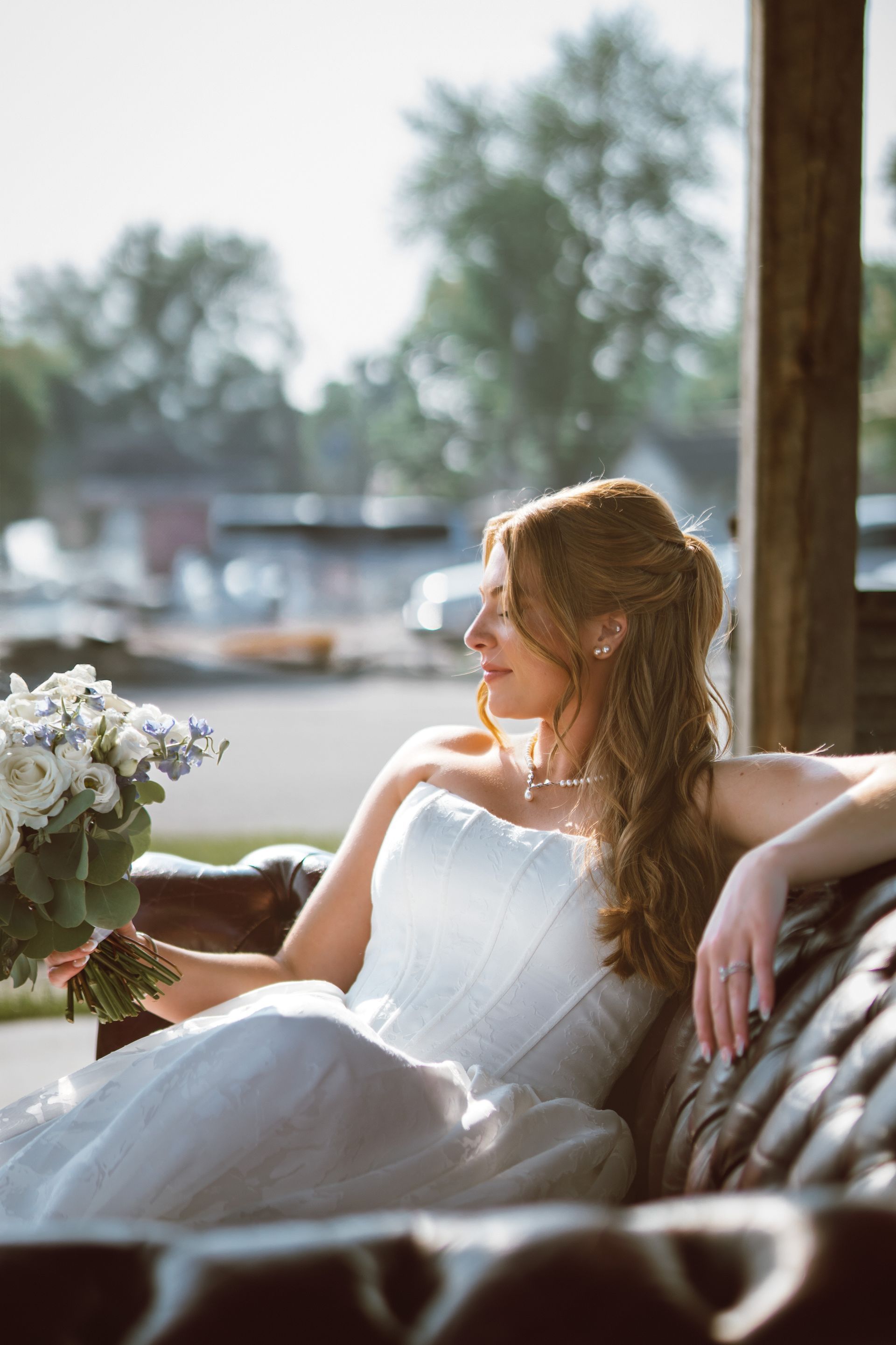 Bride in a white strapless gown sits on a leather couch, holding a bouquet. Sunlight illuminates her.