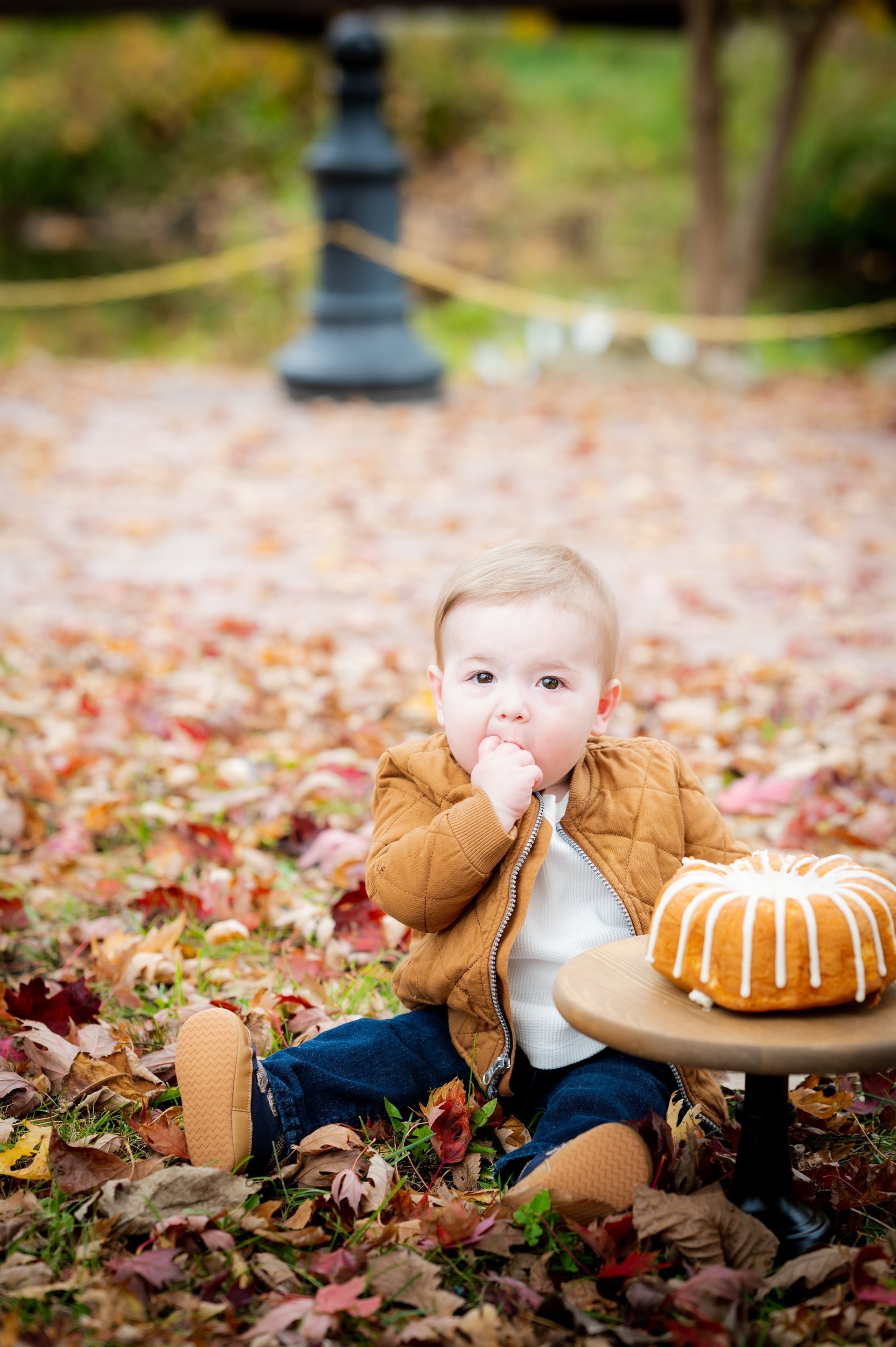 Baby sits on fall leaves, eating a finger, next to a bundt cake on a small table.