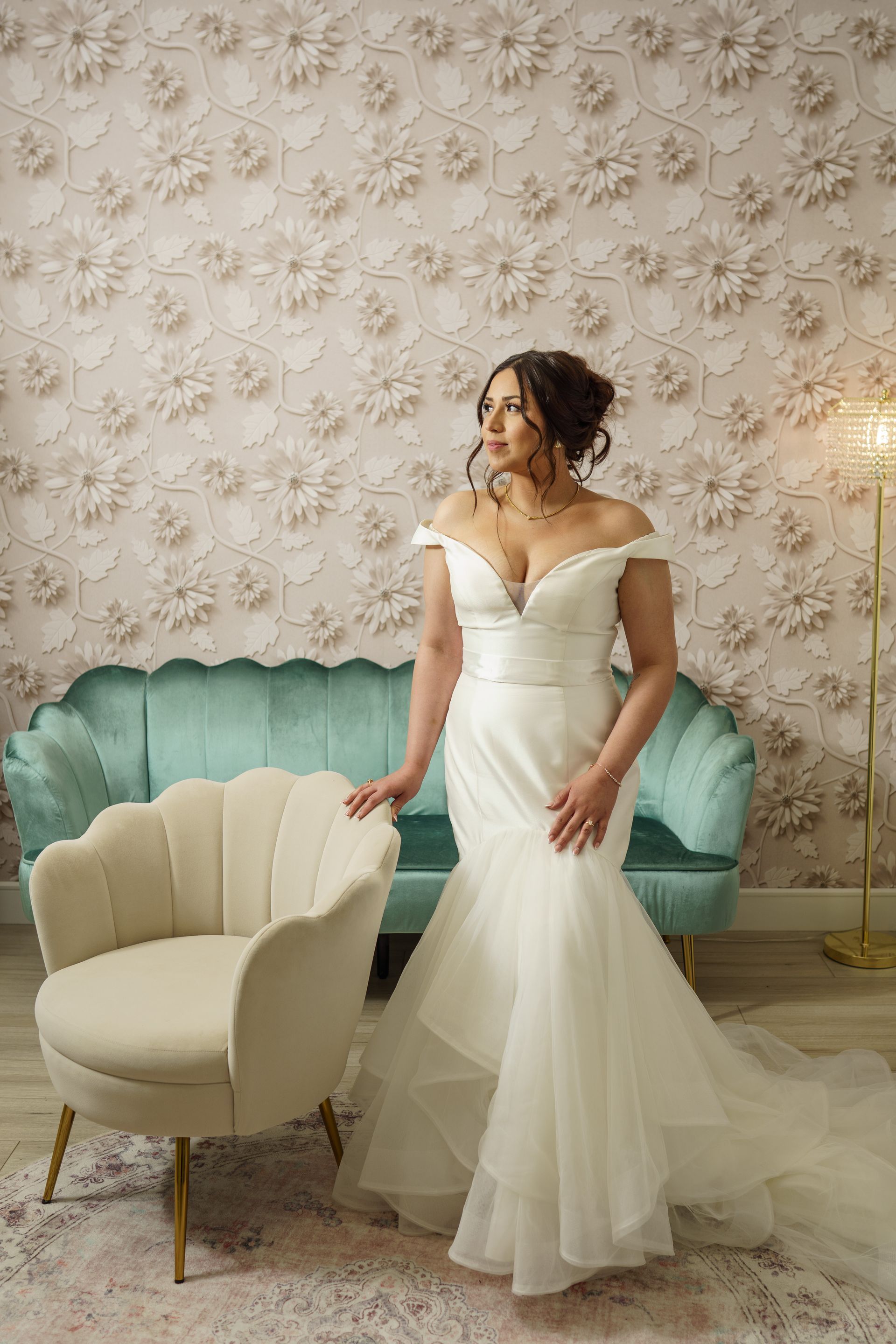 Woman in a white wedding dress, posing indoors. Leaning against a chair and looking to the side. Teal sofa and floral wall.